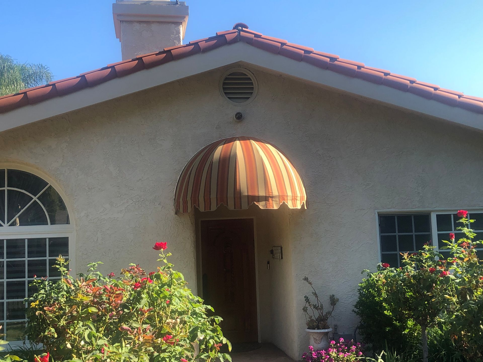 Single-story stucco house with red roof, arched windows, and striped awning over the front door
