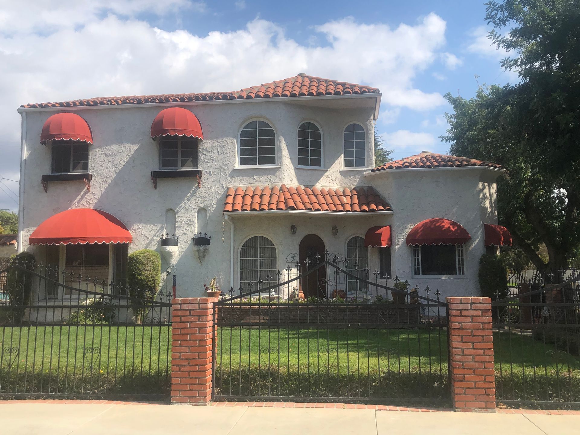 Two-story white stucco house with red awnings and a tiled roof behind a front fence under a blue sky