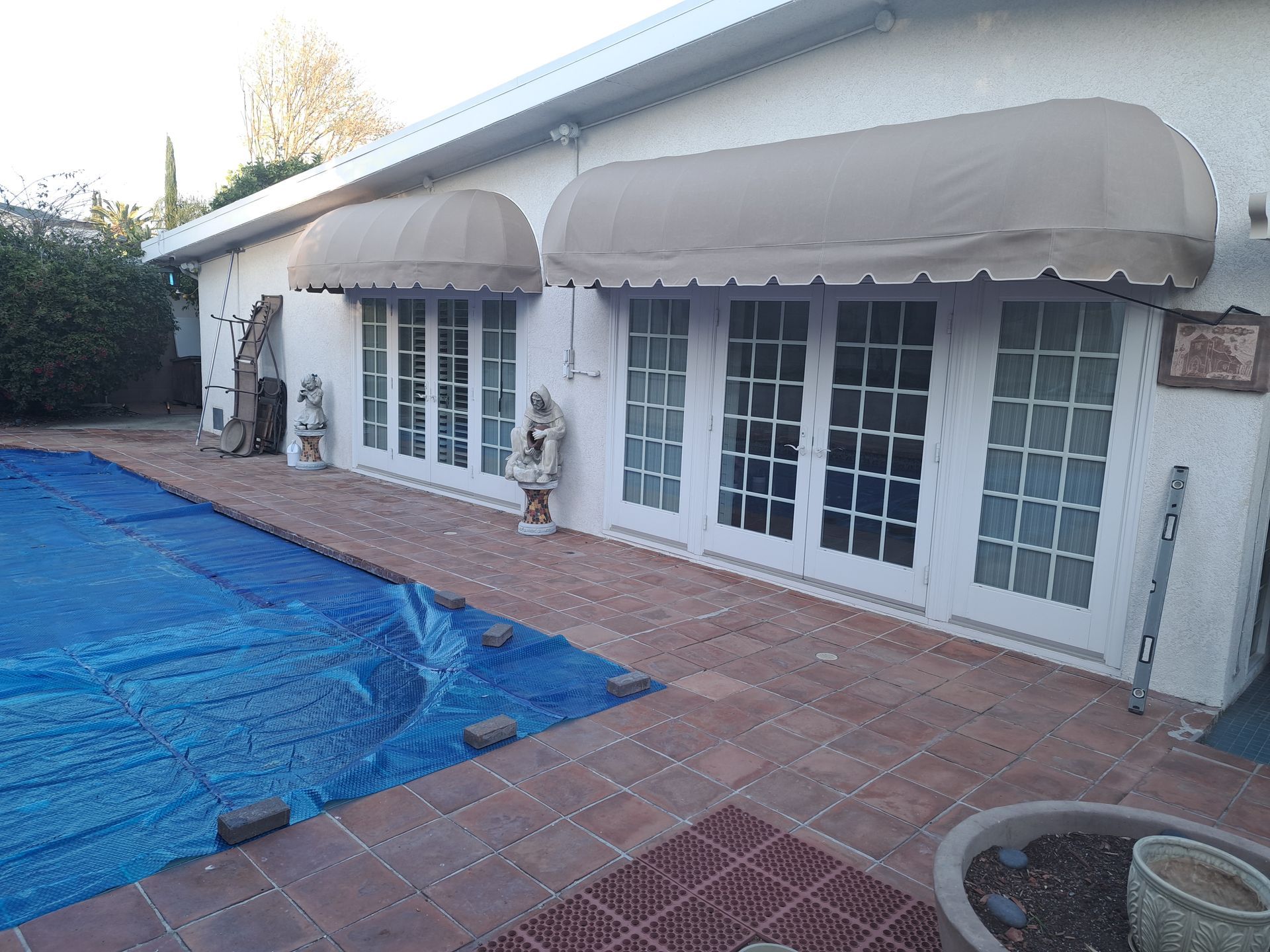 Patio with blue pool cover, brick pavers, white French doors, and striped awnings on a house exterior