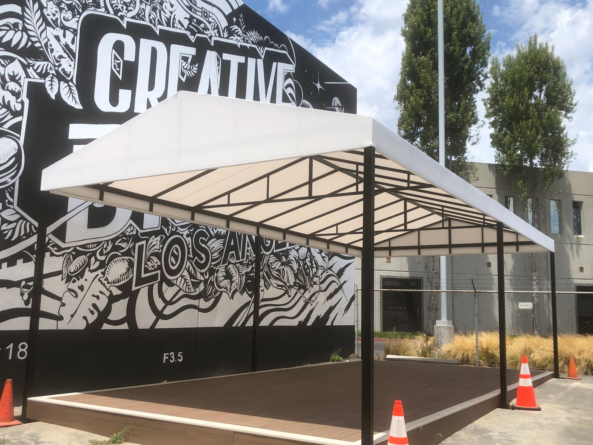White canopy tent over a wooden stage beside a mural wall with traffic cones nearby