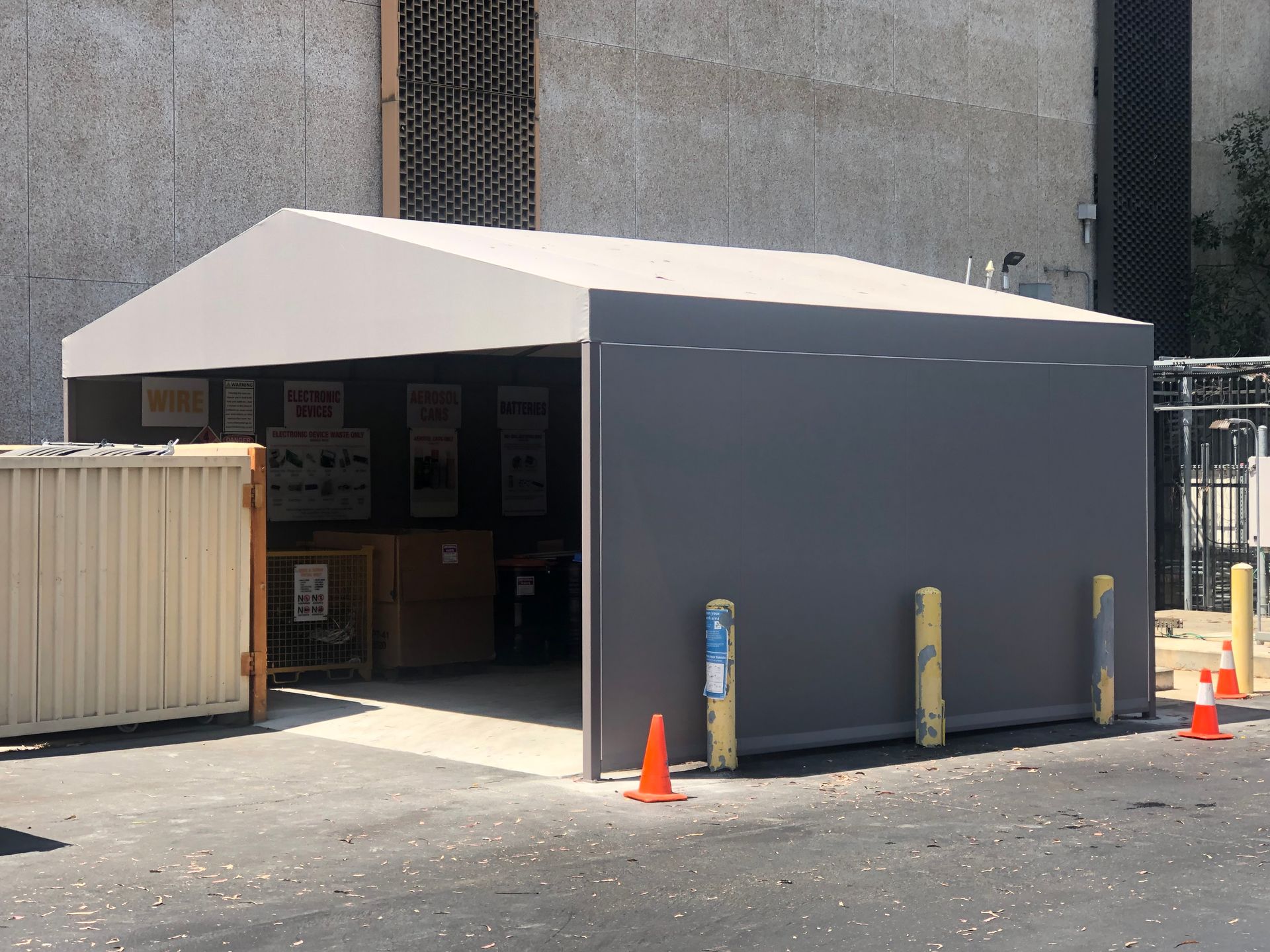 Temporary gray tent entrance outside a building, with orange traffic cones and a small container nearby