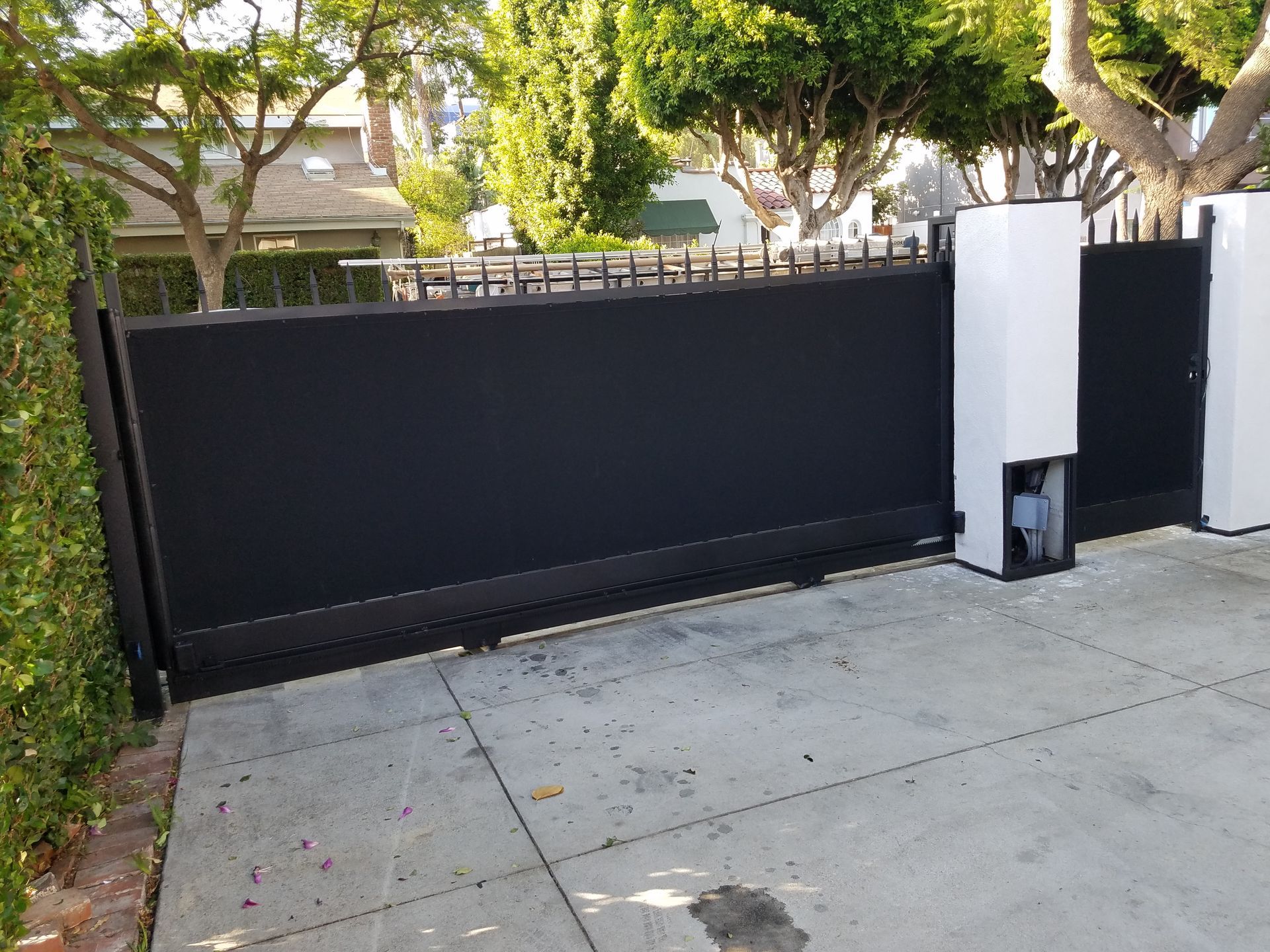 Black sliding driveway gate beside a white pillar, with a concrete driveway and trees behind it