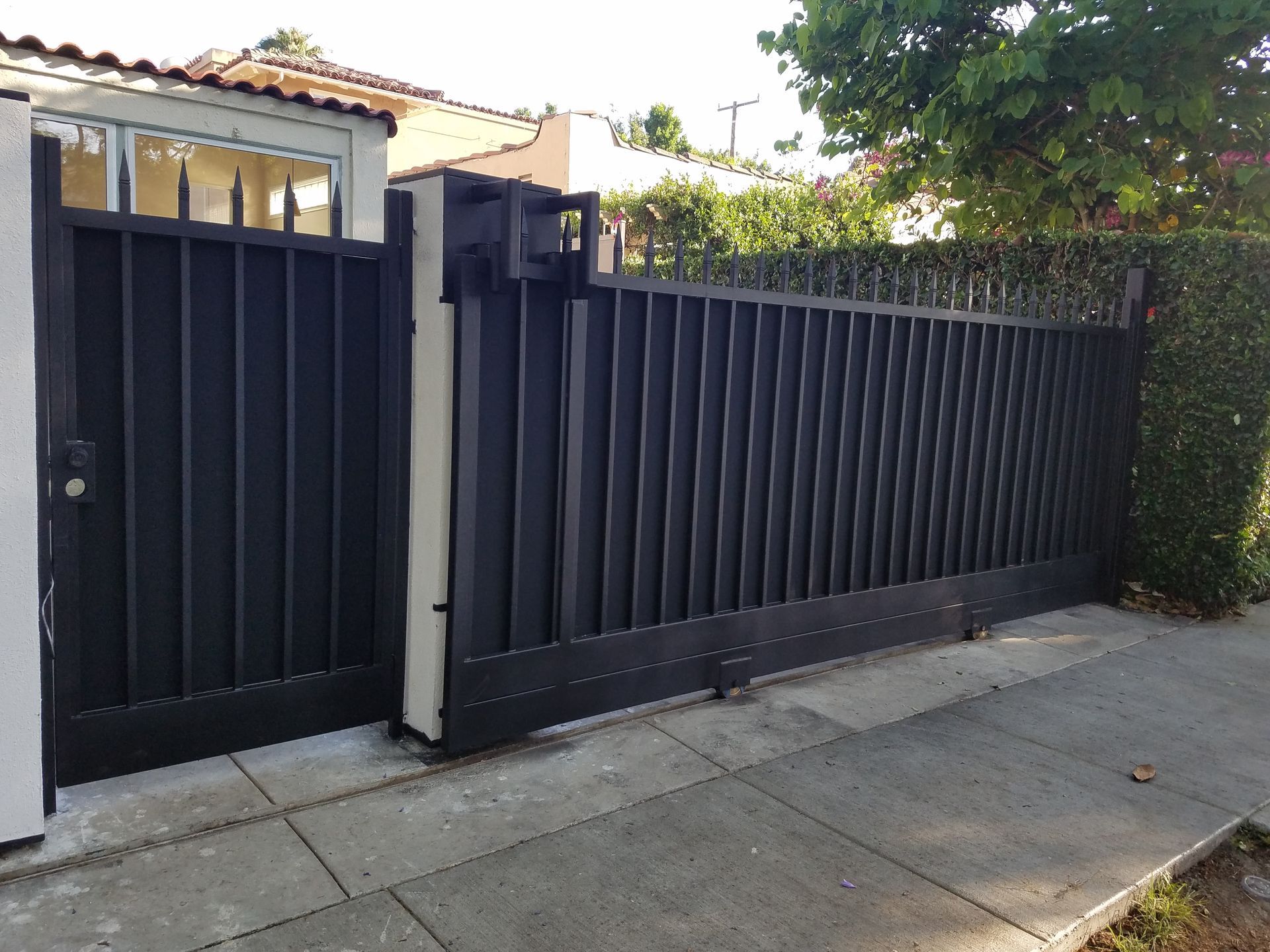 Black metal driveway gate and pedestrian door beside a sidewalk, with a house and trees behind.