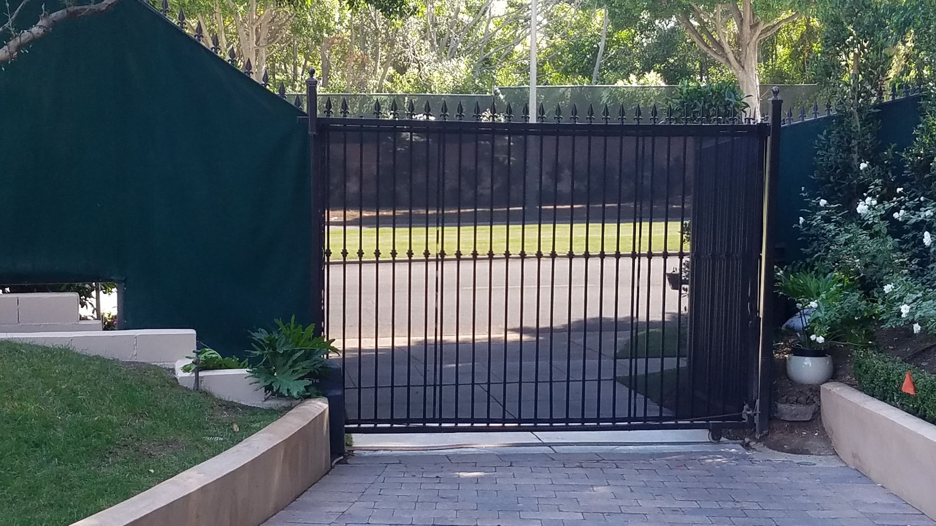 Black metal gate at the end of a paved driveway, bordered by plants and a driveway wall.