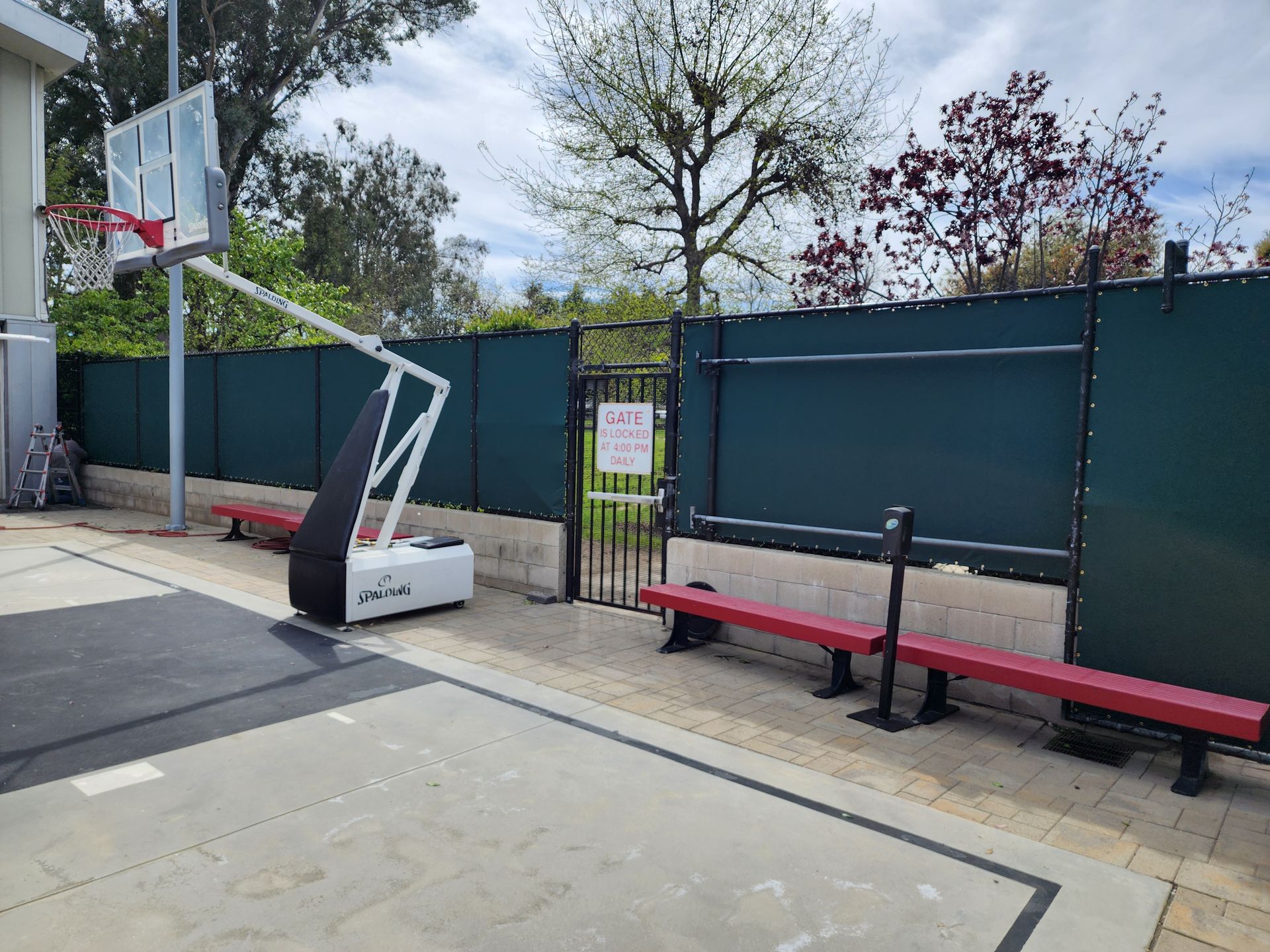 Outdoor basketball court with a hoop, red bench, and tall green fence under cloudy skies