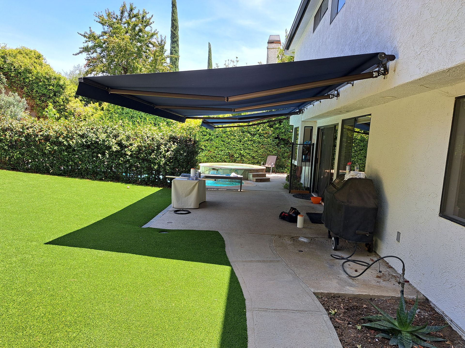 Backyard patio with black awning, white house, green lawn, and outdoor furniture.