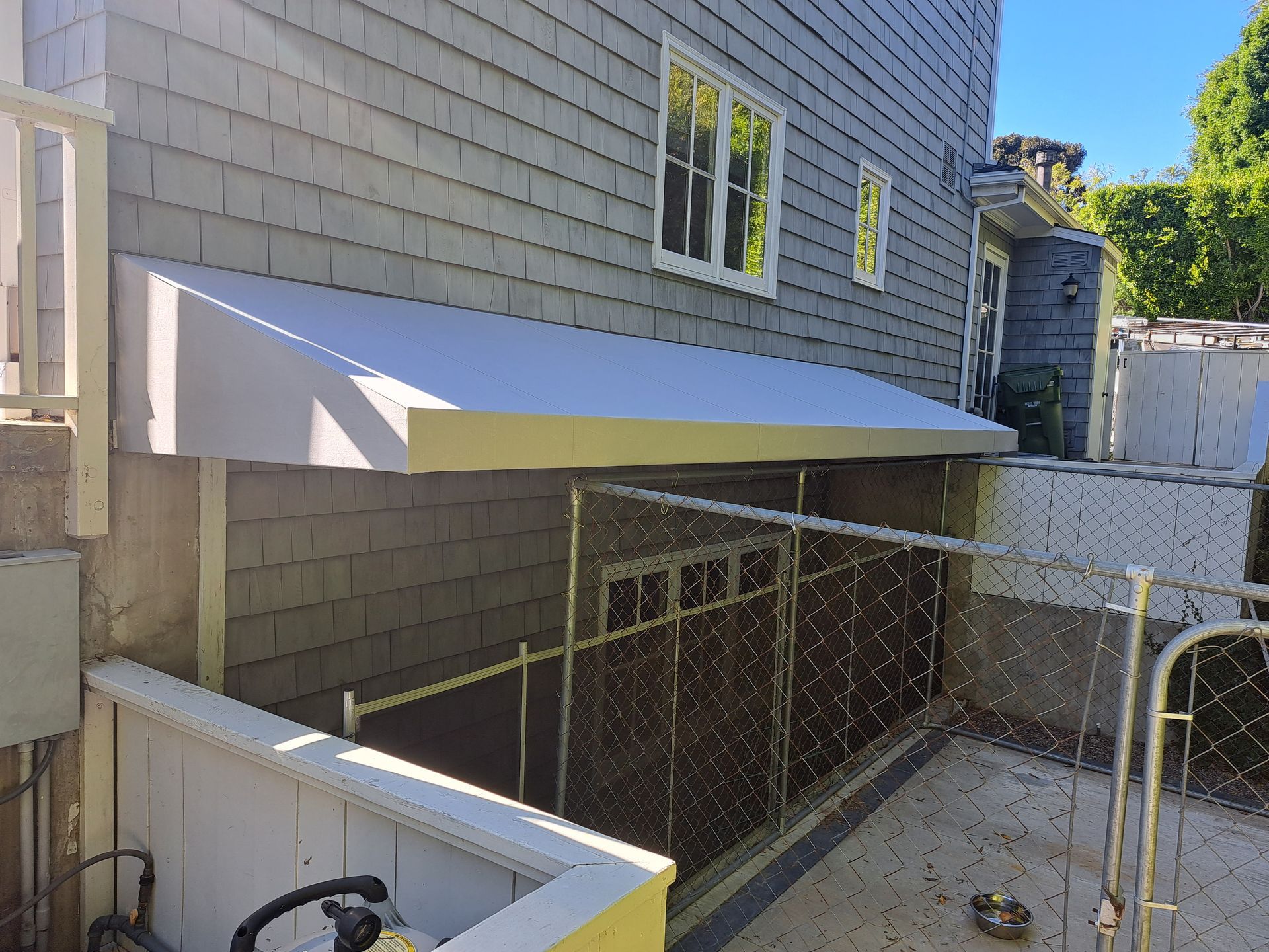 Backyard side entrance with white awning, chain-link fence, and basement windows beside a gray house