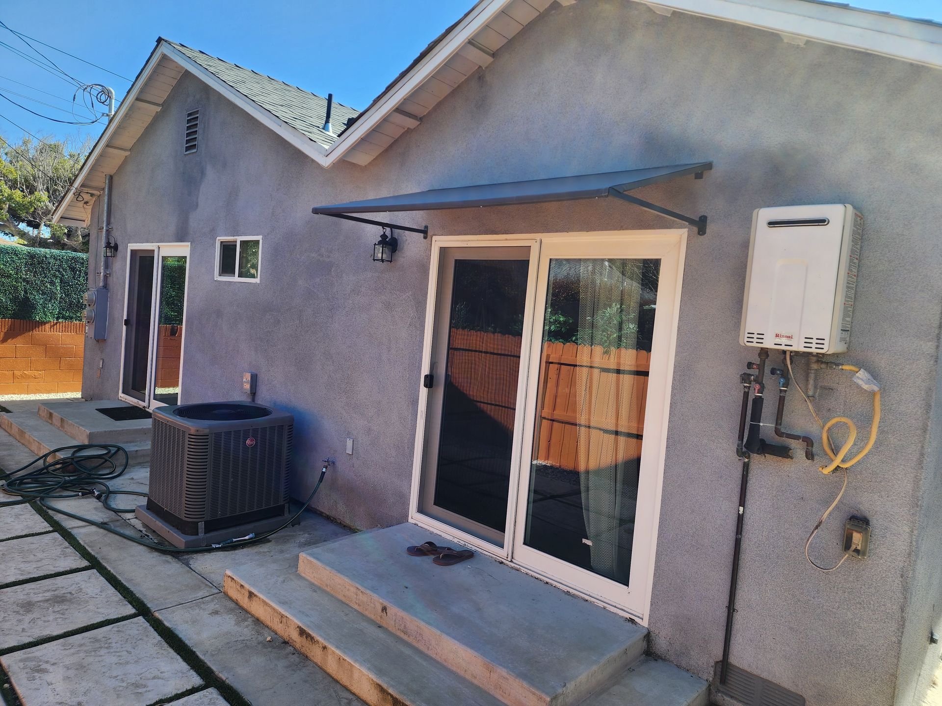 Small gray house exterior with sliding glass doors, steps, and an air conditioner unit on a paved patio