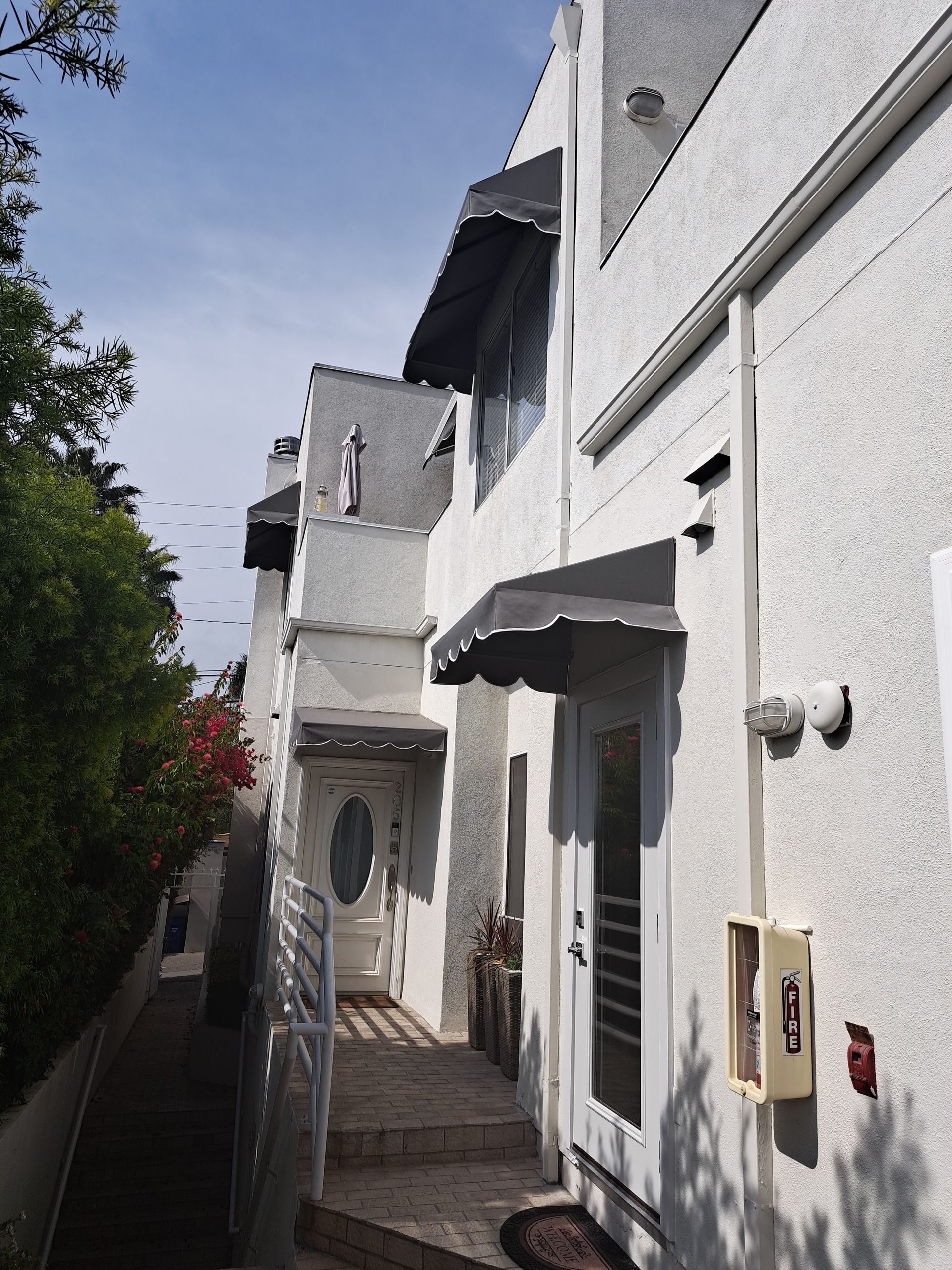 Narrow white stucco alley with doorways and awnings beside greenery under bright sunlight