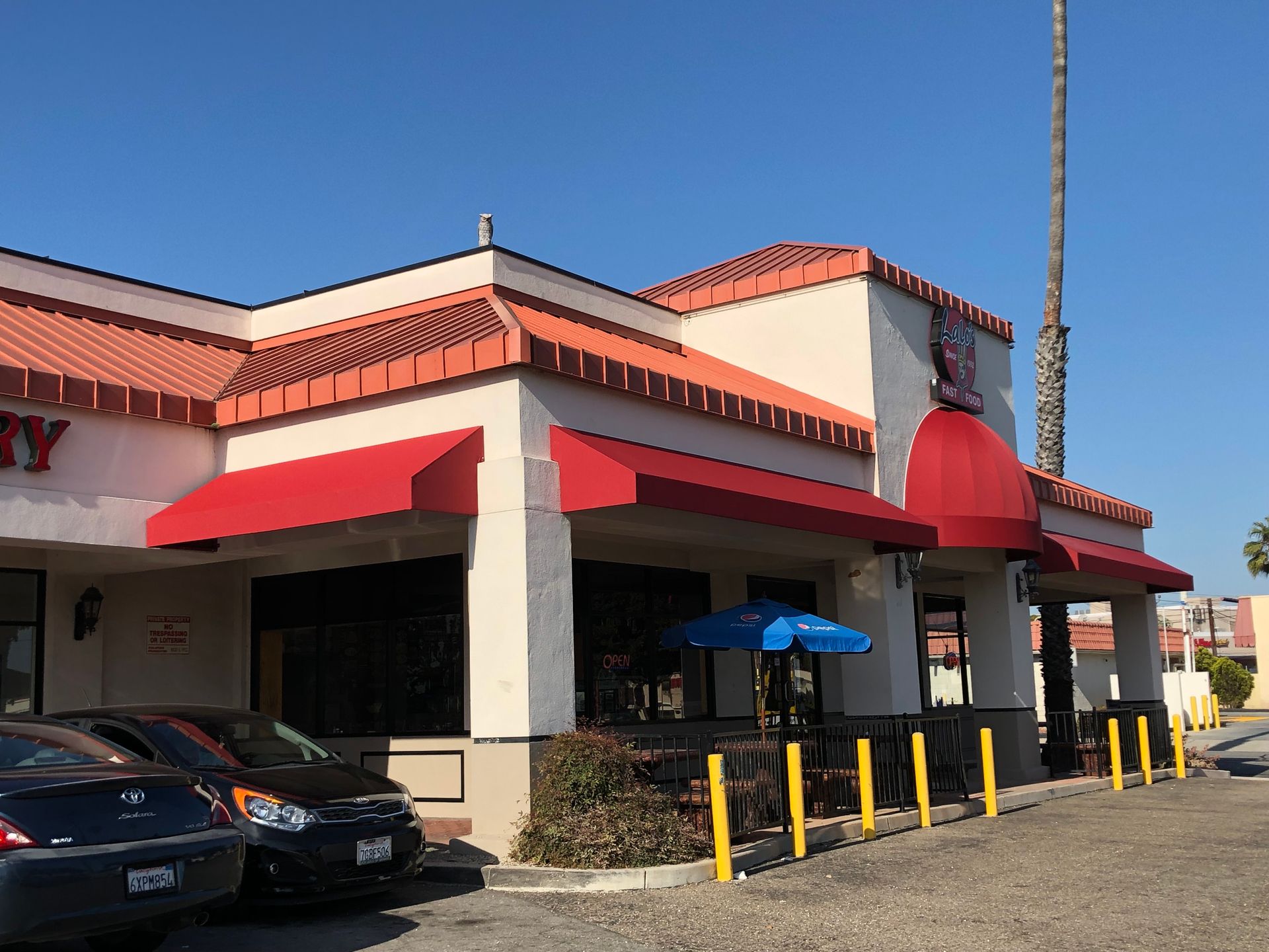 Restaurant exterior with red awnings, tiled roof, parked cars, and blue patio umbrellas