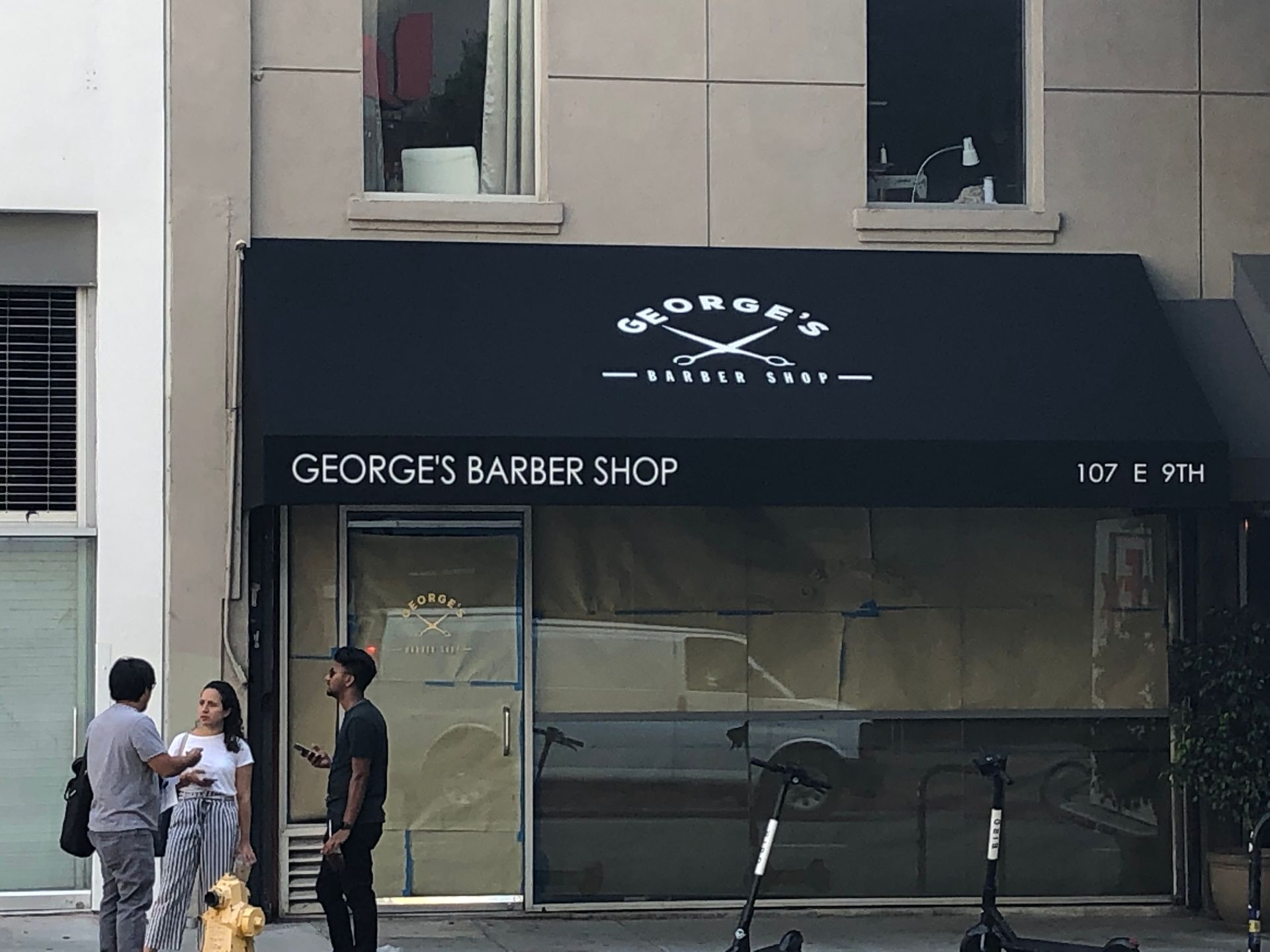 People standing outside Georges Barber Shop with a striped awning and two e-scooters on the sidewalk