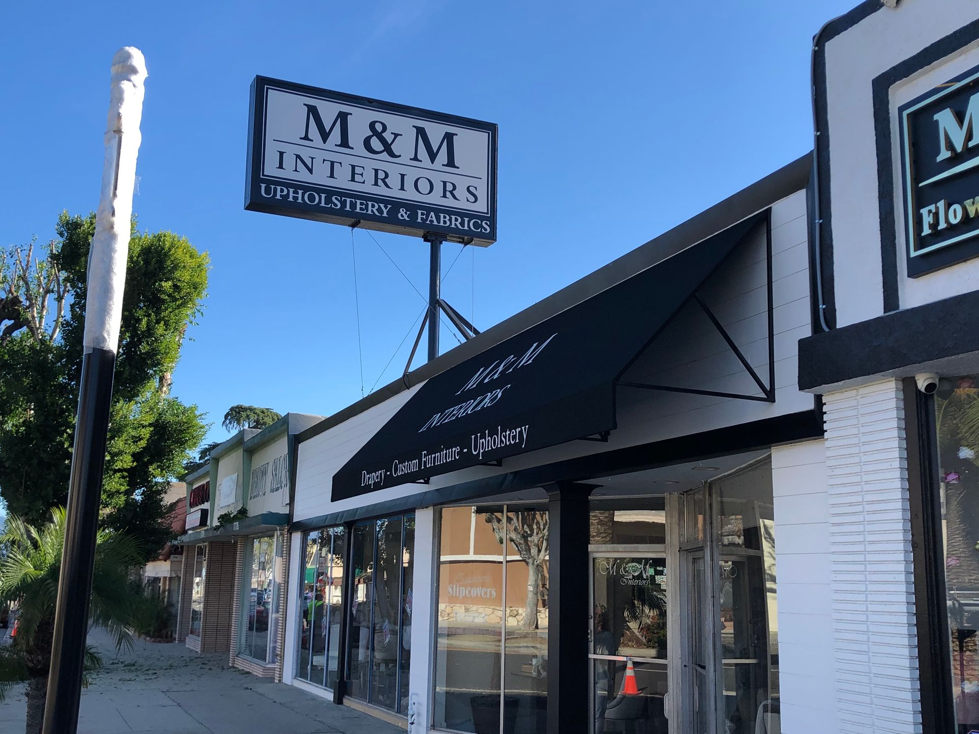 Storefronts along a sunny street, including an M&M Interiors sign and black awnings.