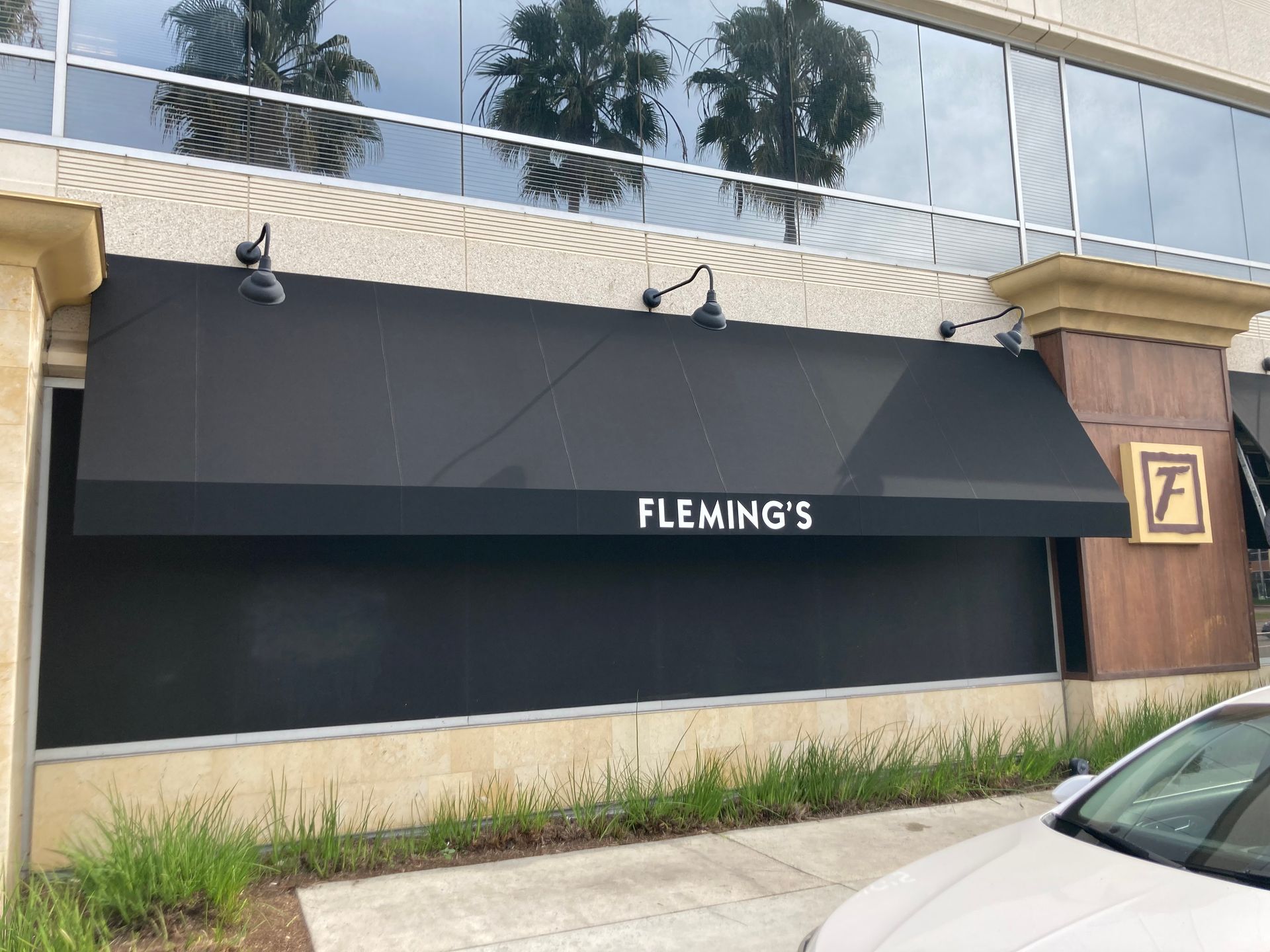 Storefront with dark awning labeled “FLEMING’S” on a beige building, with a car at the curb.