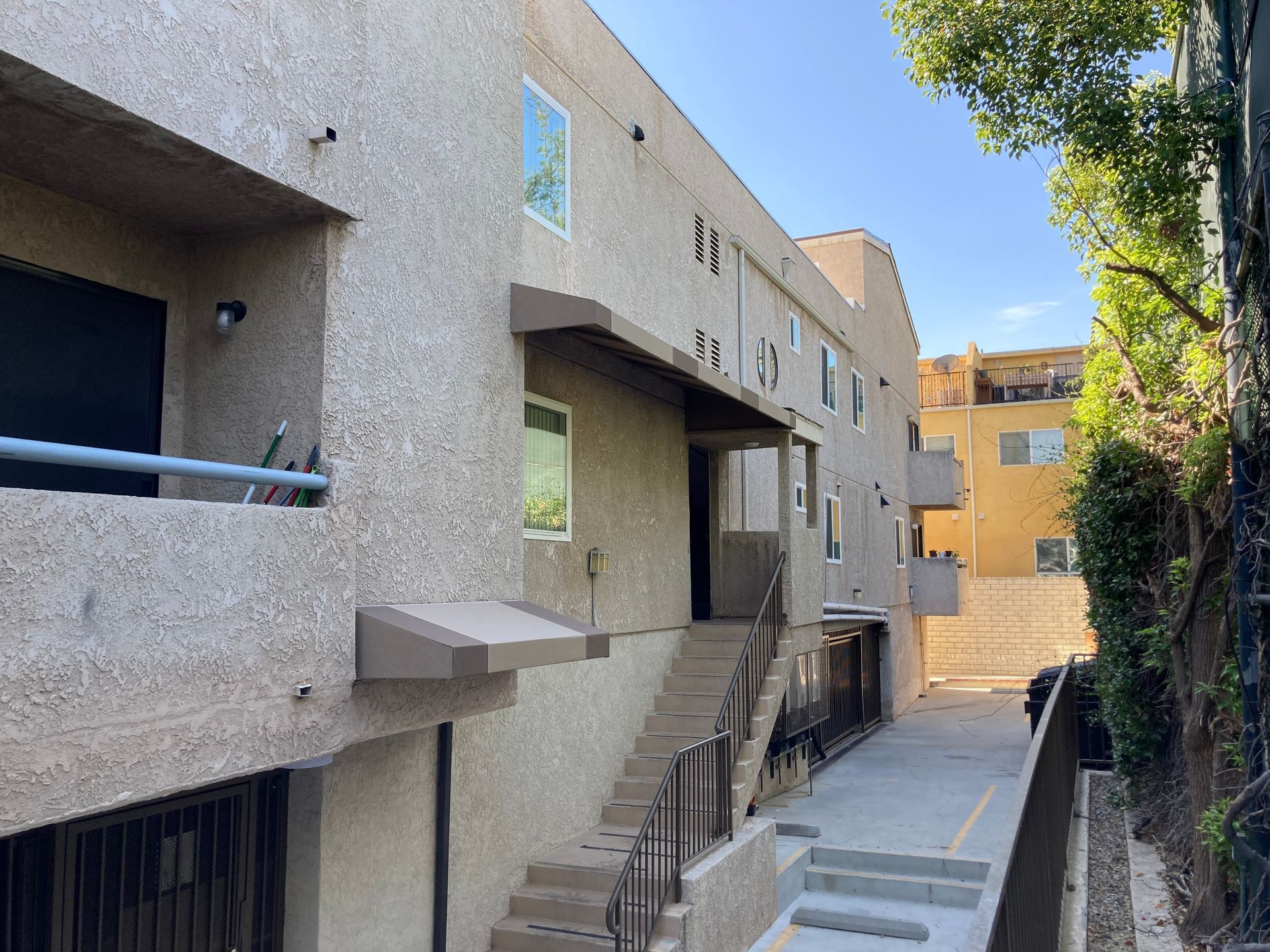 Exterior walkway beside beige stucco apartment buildings, with stairs and a narrow path under a blue sky.