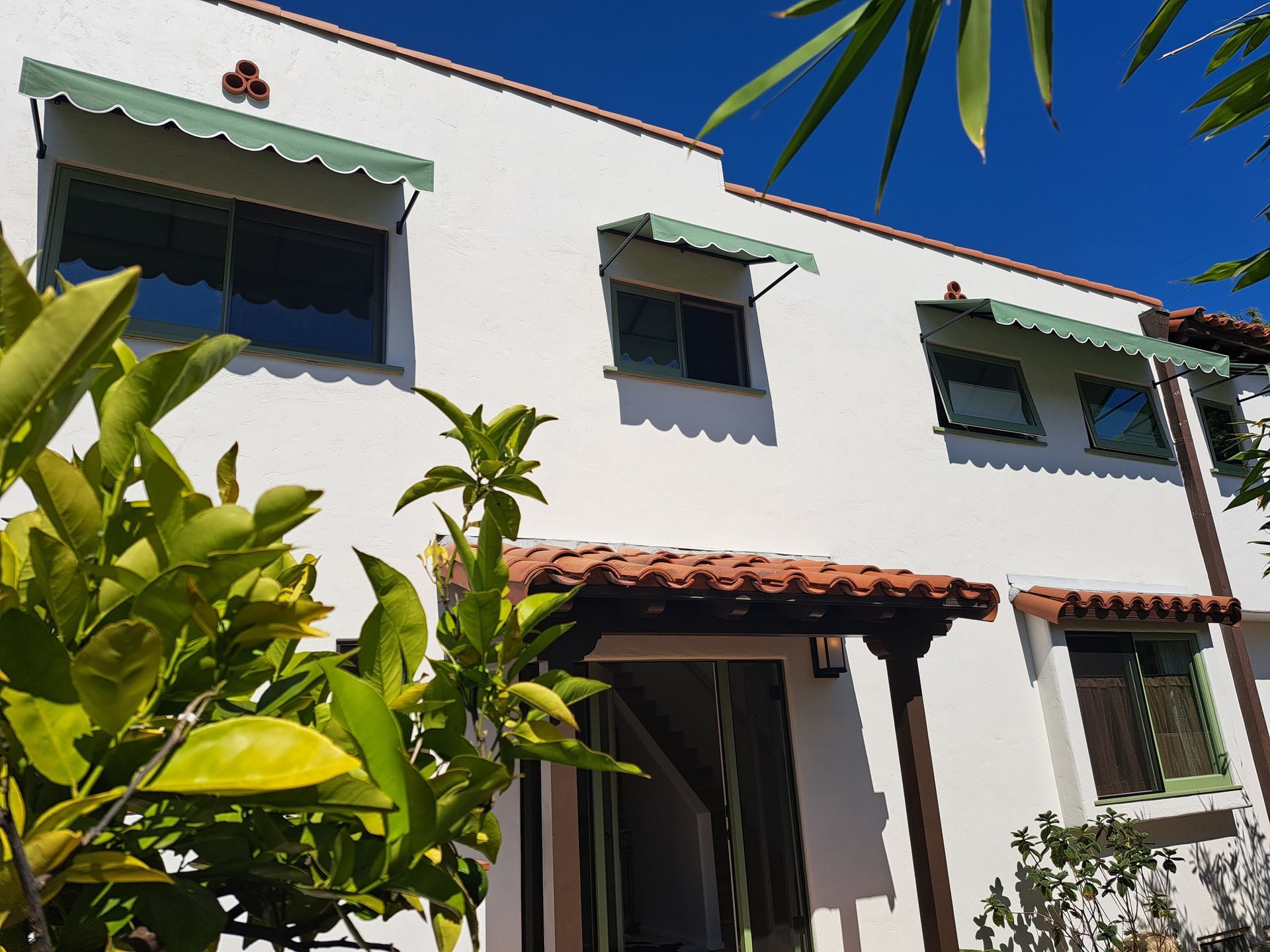 White two-story house with green awnings and a red-tiled porch, framed by leafy plants under a blue sky.