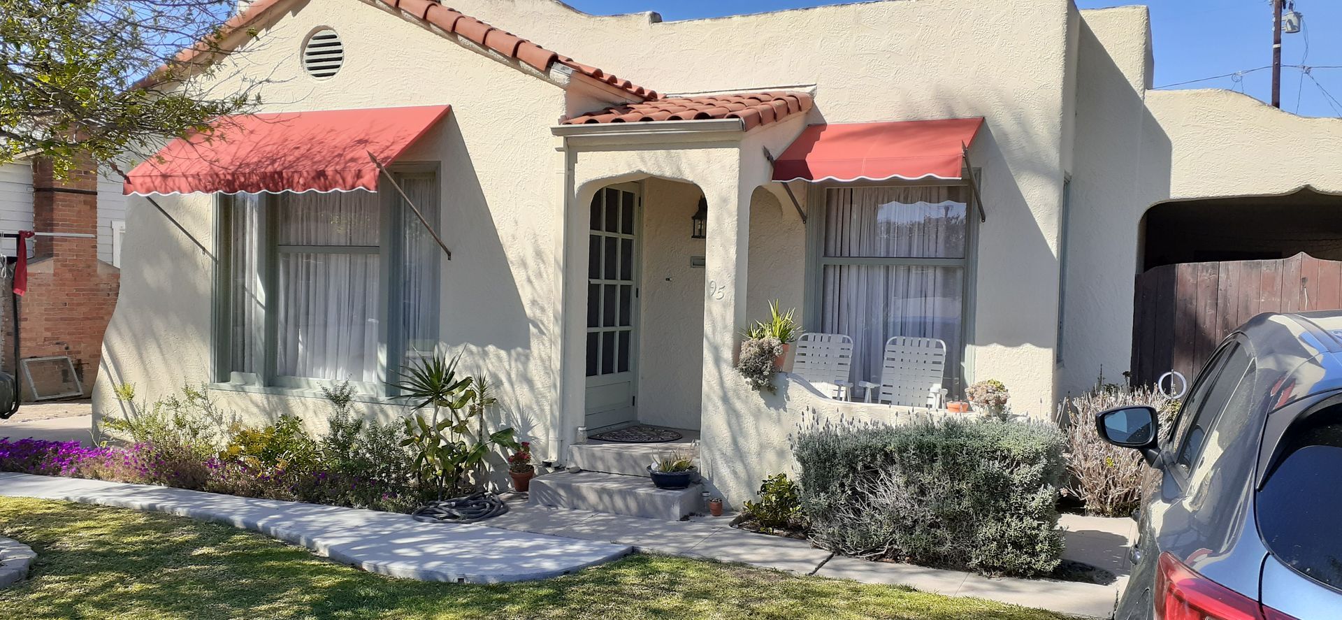 Small stucco house with red awnings, white curtains, and a red car parked at the curb