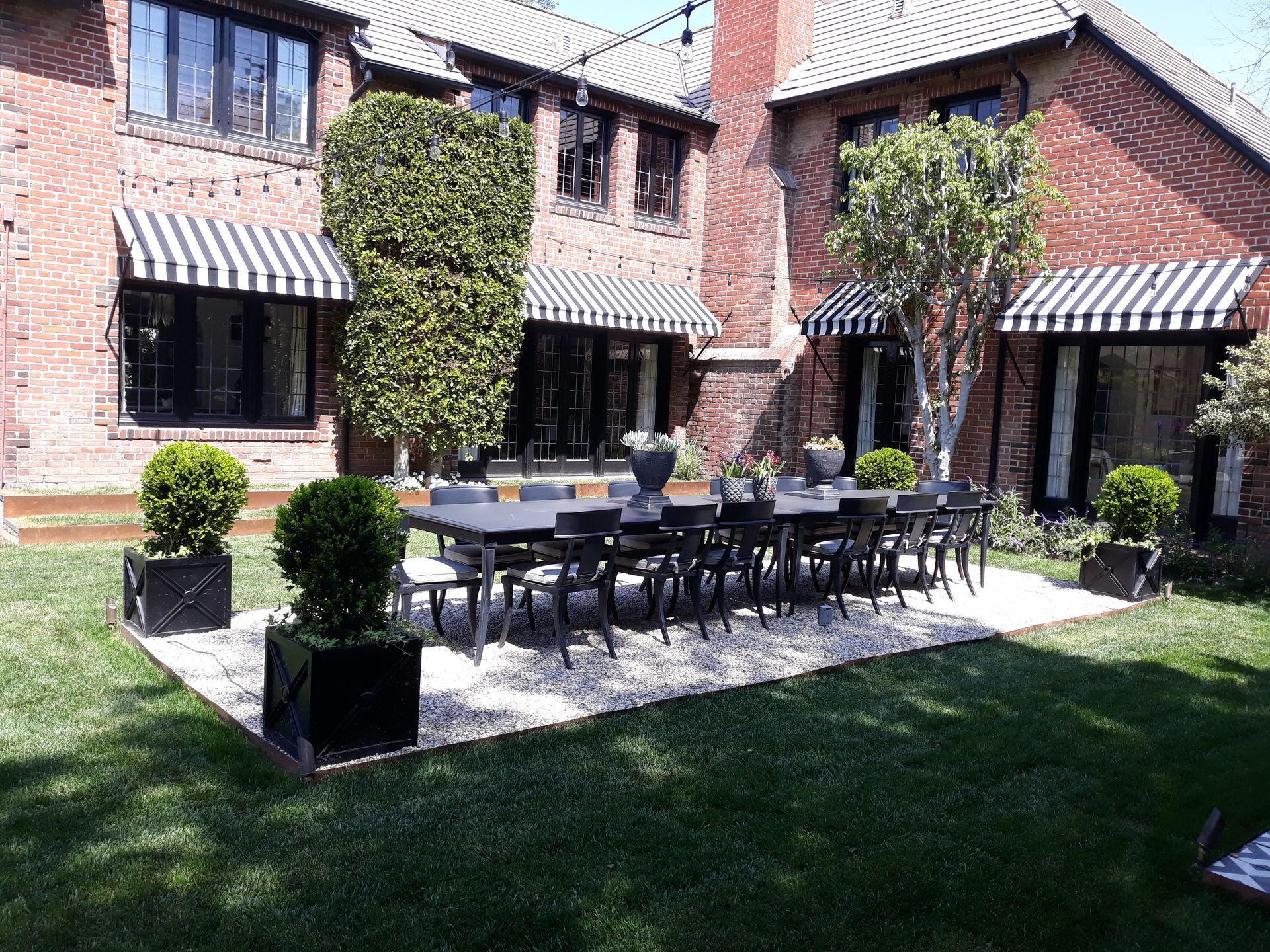 Brick courtyard patio with black outdoor furniture, striped awnings, and potted plants beside a lawn