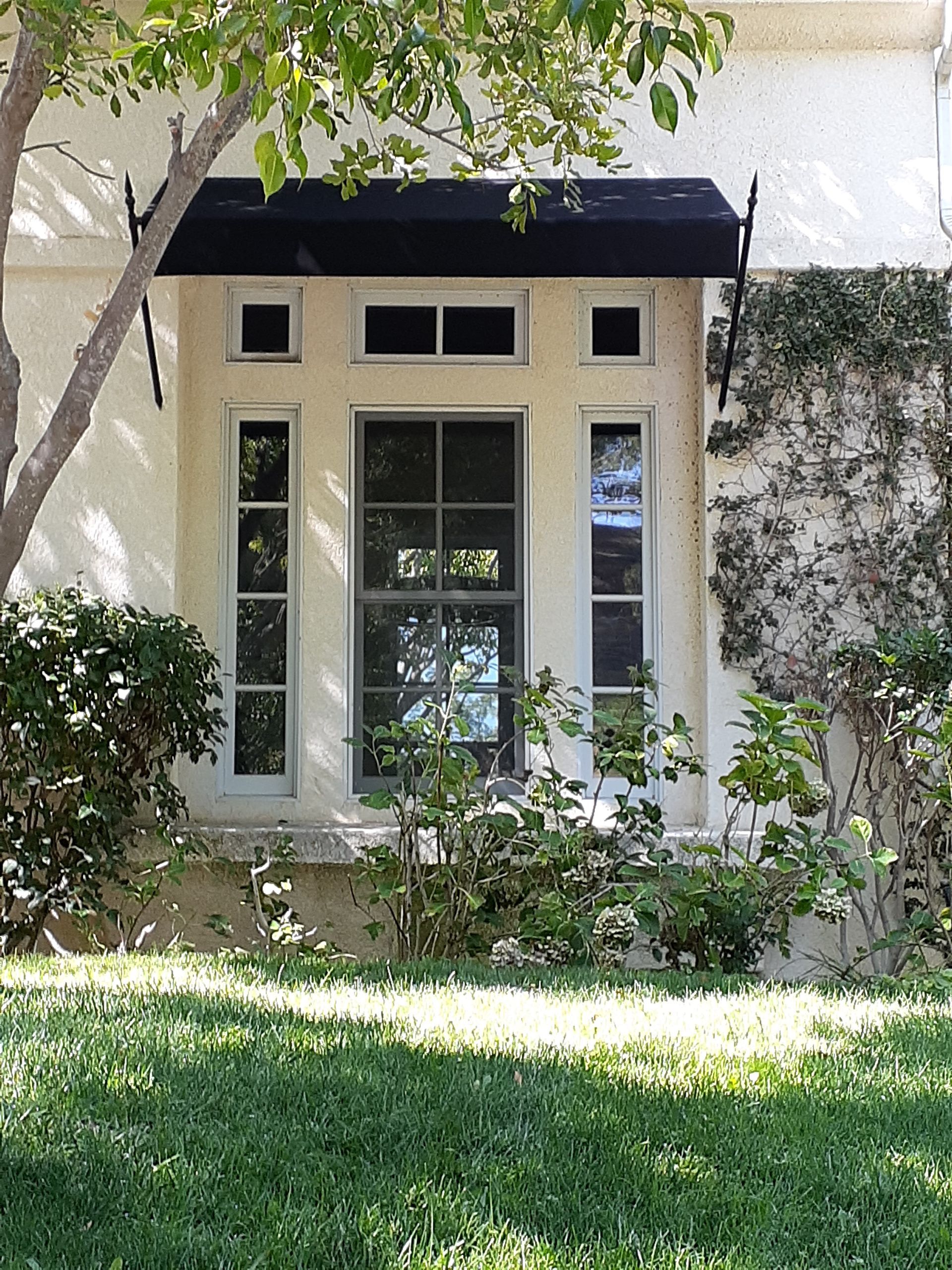 Front of a house with a black awning, cream trim, and windows framed by greenery.