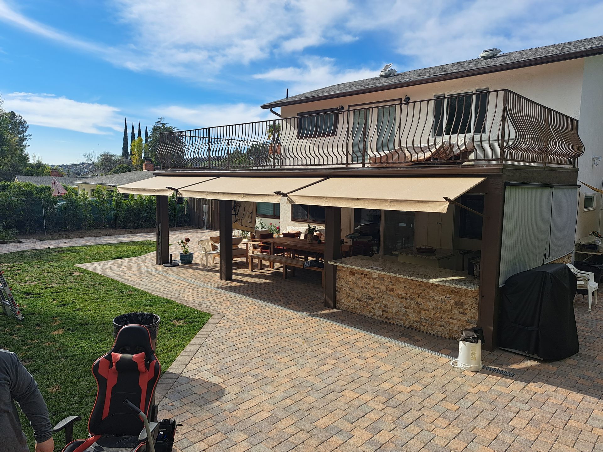 Backyard patio with a tan canopy, stone pavers, picnic table, lawn, and a two-story house under a blue sky