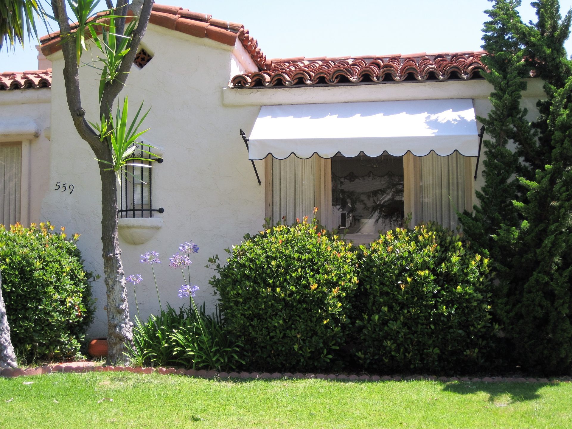 White stucco house with red tile roof, awning, and lush green shrubs in the front yard
