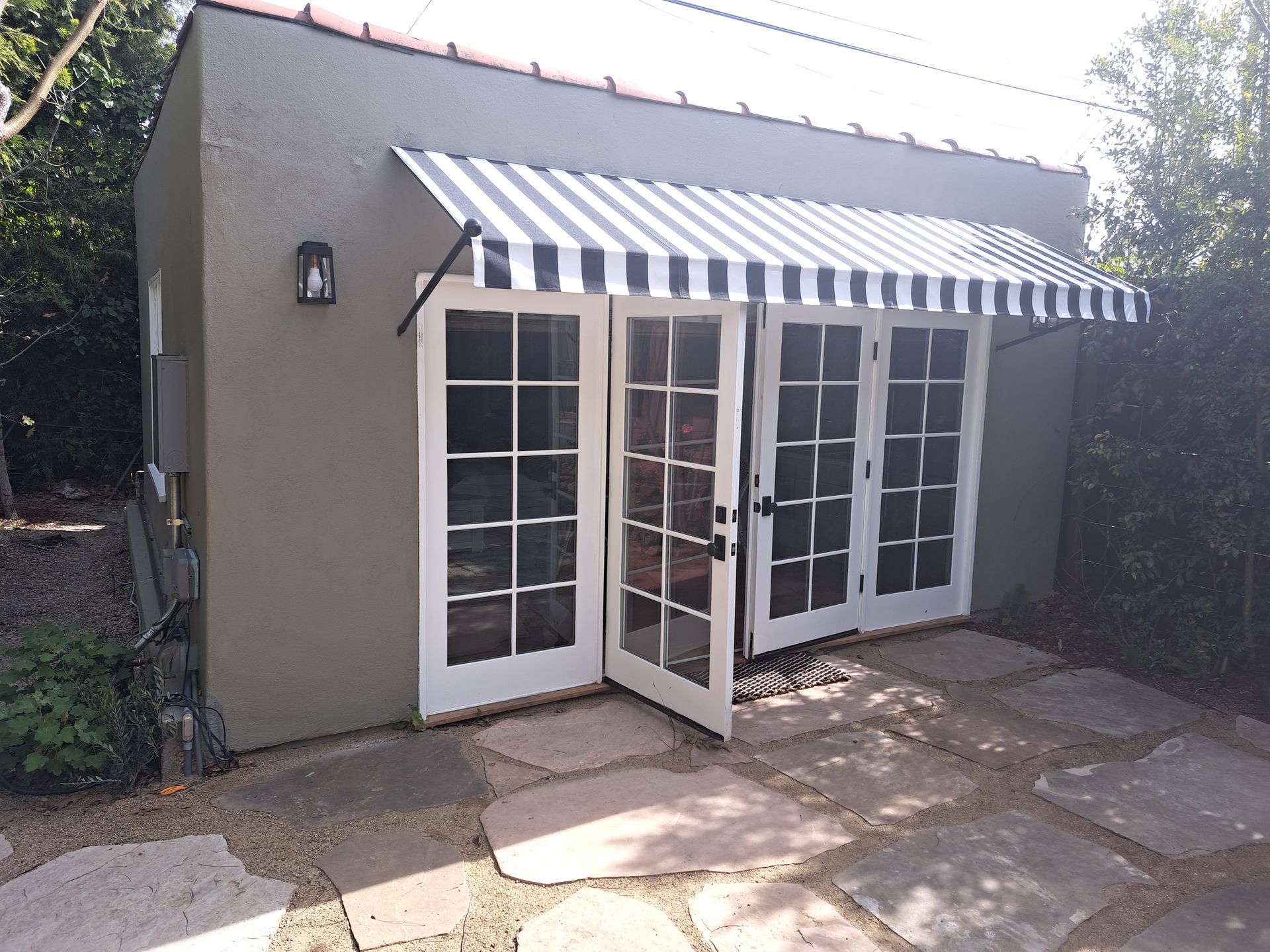 Small patio with a beige building, striped awning, and open French doors in sunlight