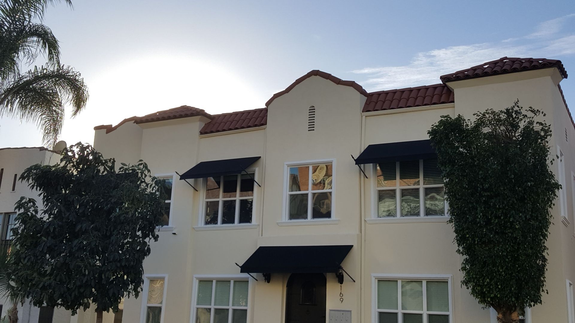 A cream-colored apartment building with red tile roofs, black awnings, and palm trees on either side.