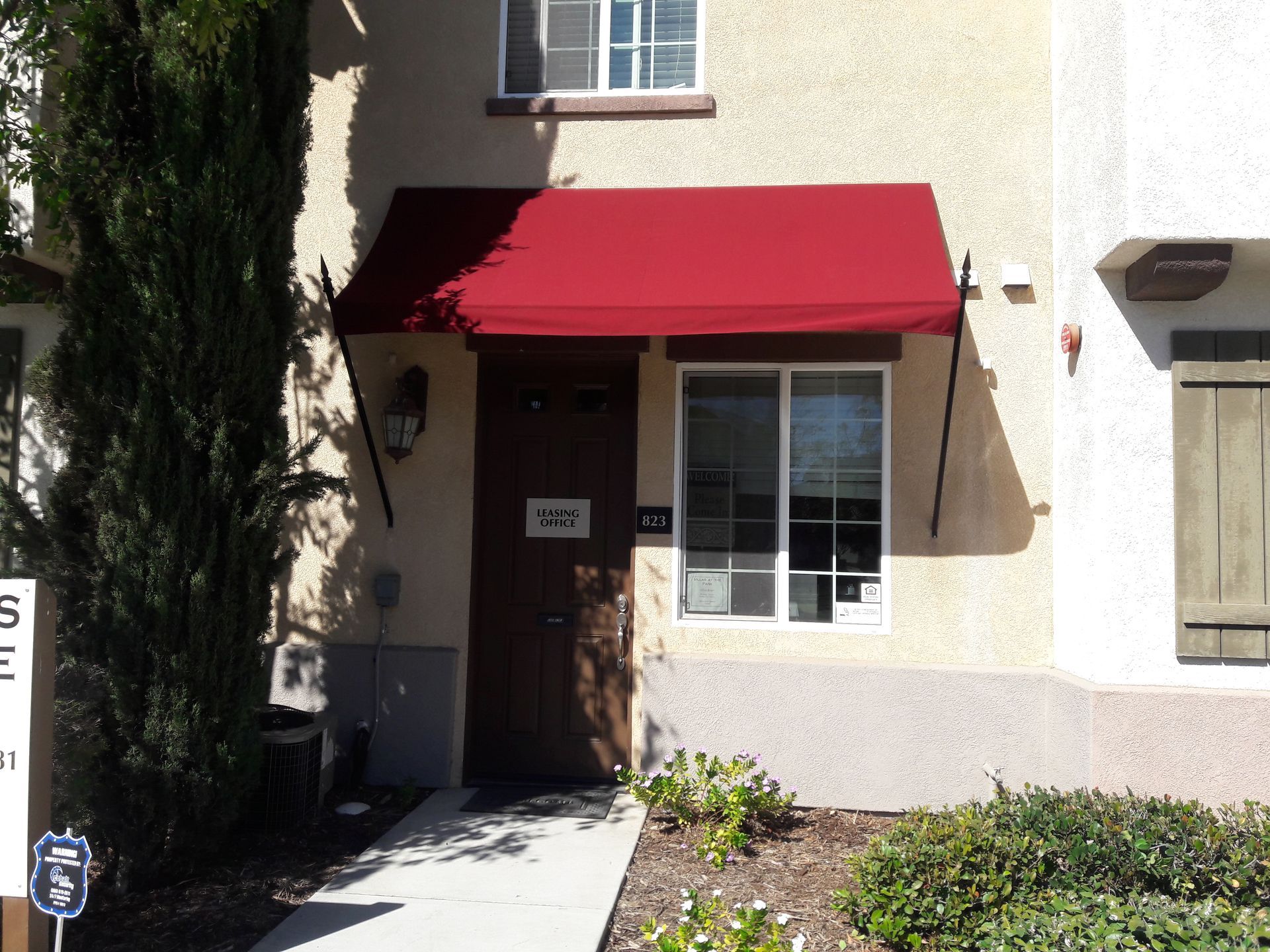 Building entrance with a red awning and a white-framed door, surrounded by stucco walls and shrubs.
