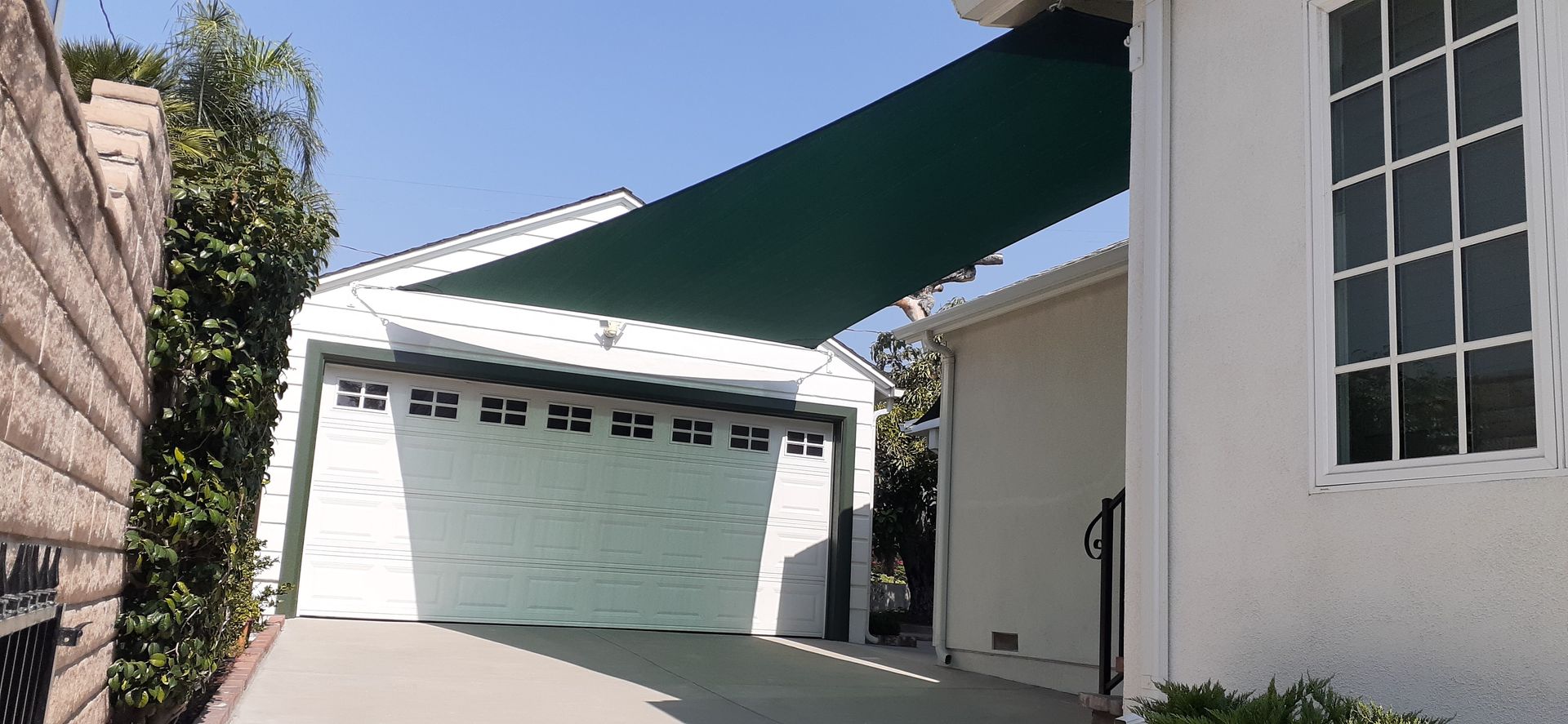 White house with green garage door and awning, viewed from a driveway beside stone walls.