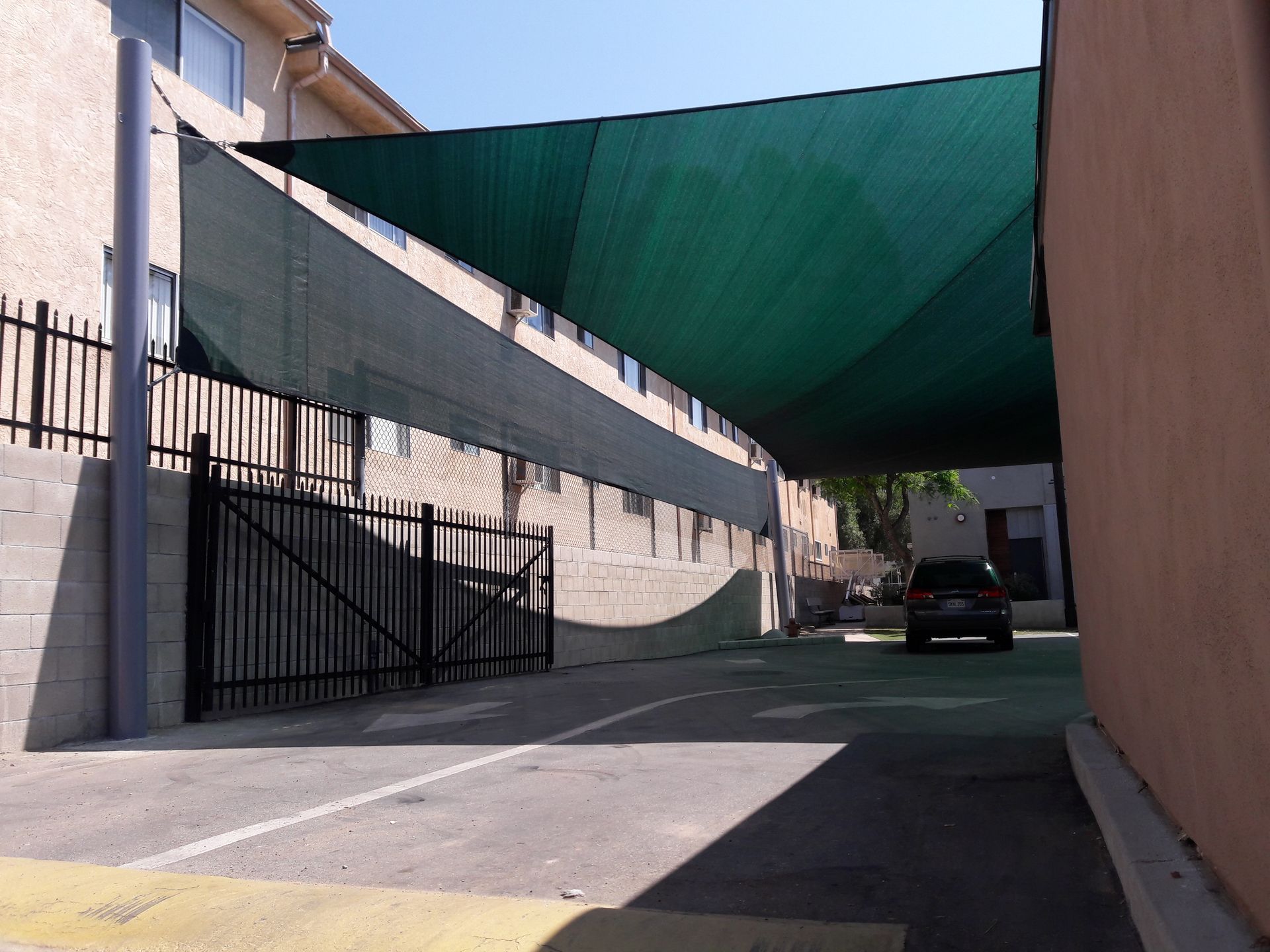 Narrow alleyway with green shade sails overhead, black gate, and a parked car in the distance