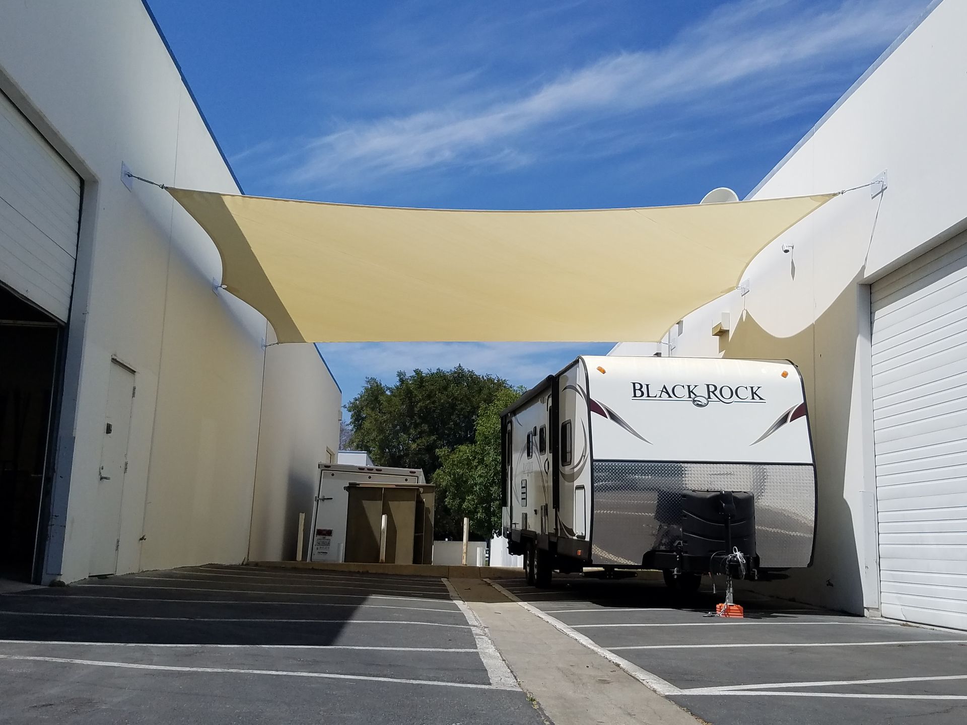 White industrial alley with a camper trailer parked under a tan shade sail on a bright day