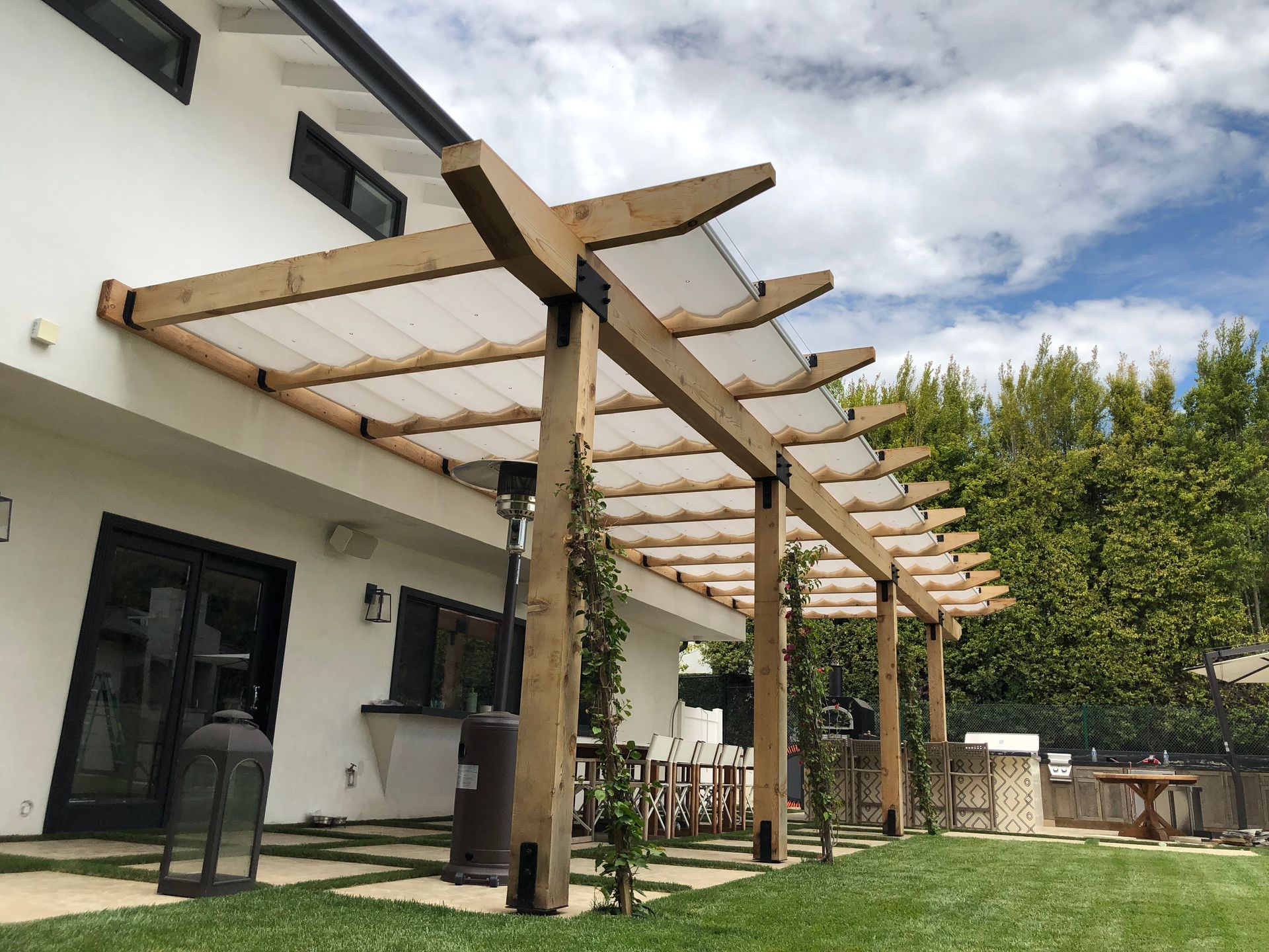 Patio with wooden pergola and seating beside a white house, with trees in the background