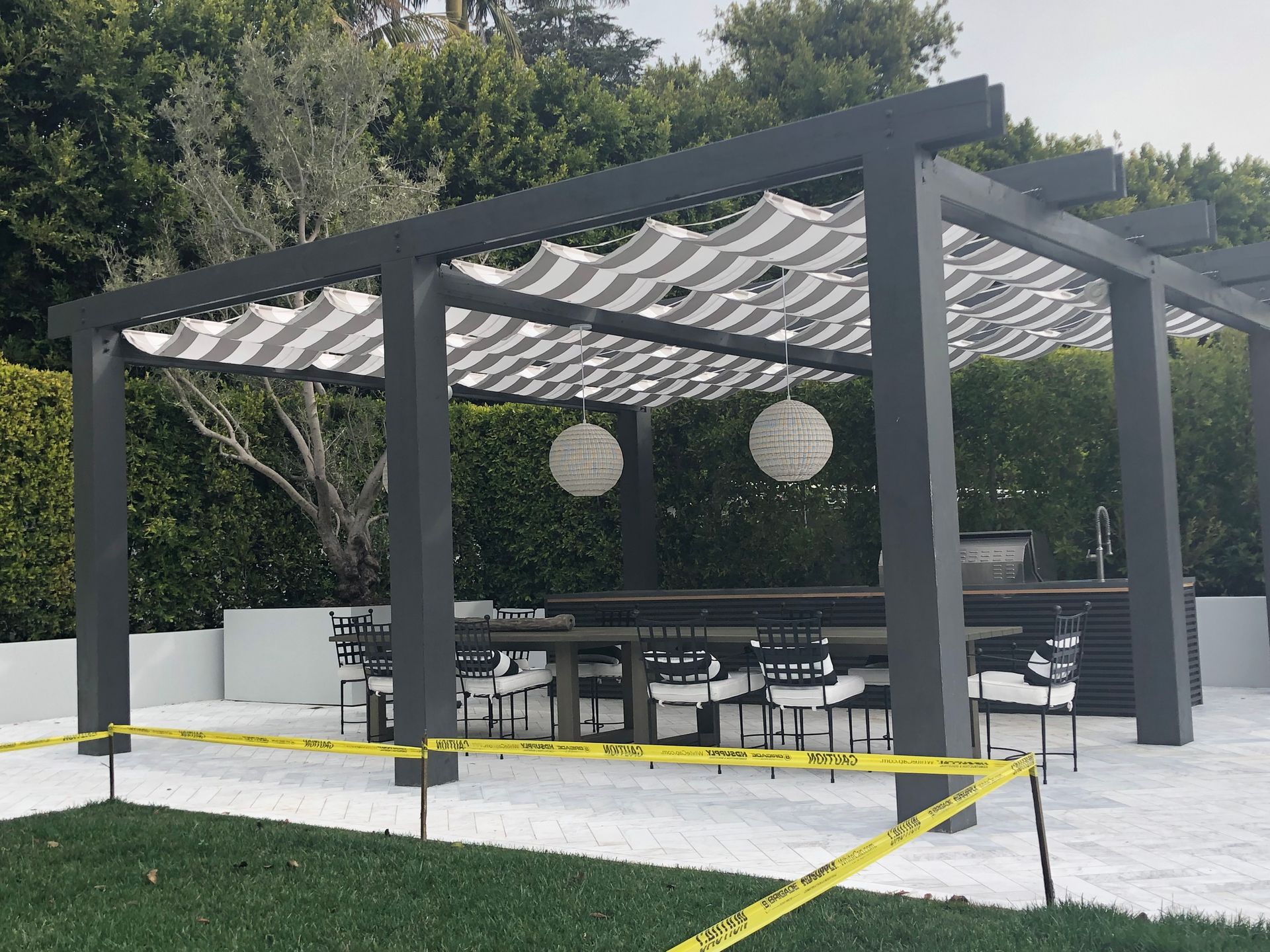 Modern black pergola with outdoor seating on a white patio, surrounded by trees and grass.