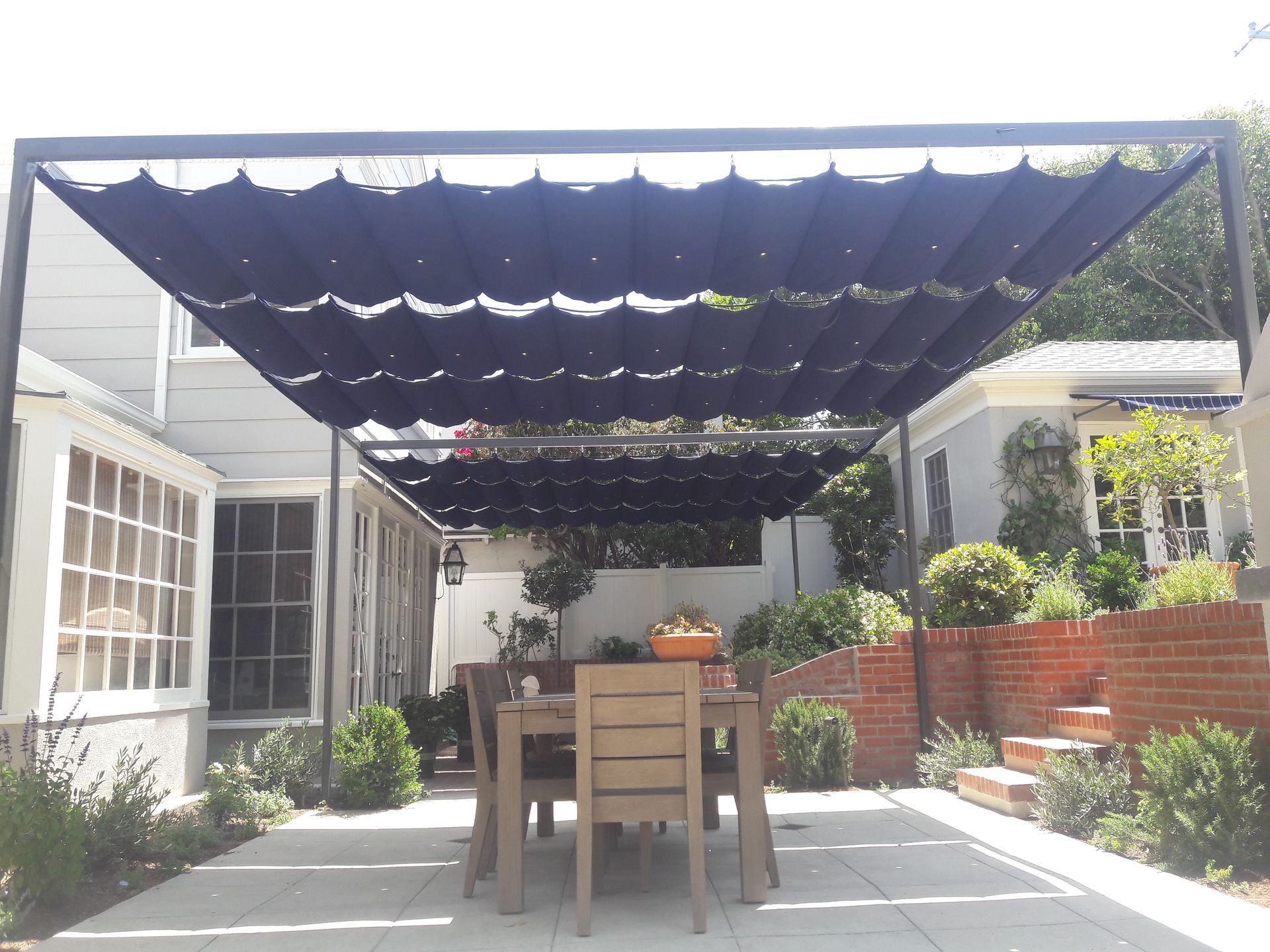 Shaded patio with wooden table and chairs under a dark canopy, surrounded by plants and brick planters