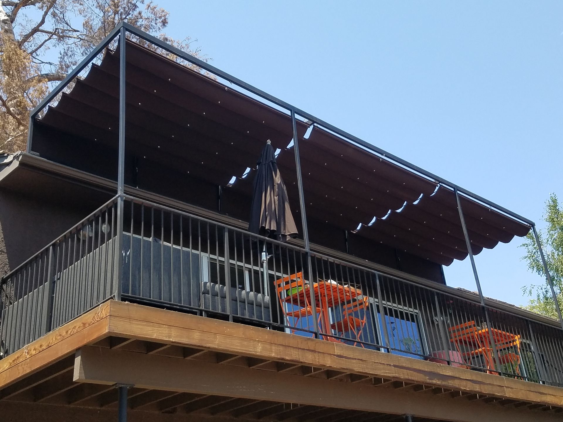 Outdoor wooden deck with a dark metal railing, red flowers, and a striped canopy under a blue sky