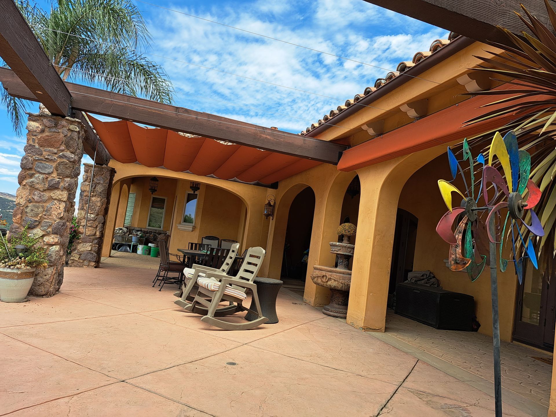 Patio with stone columns, tan walls, lounge chairs, and colorful wind spinner under a blue sky
