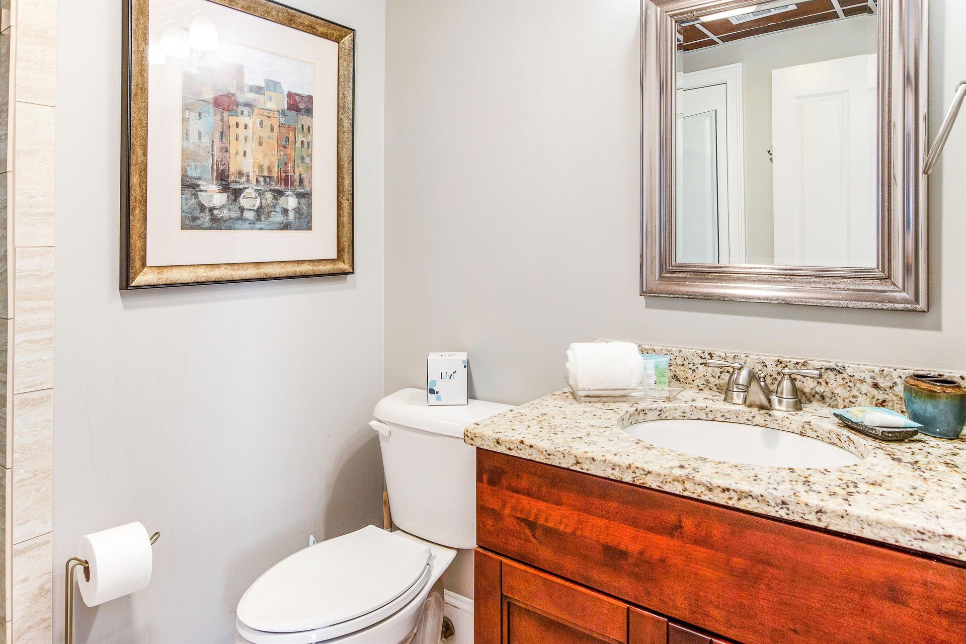 Bathroom with gray walls, wood vanity, granite countertop, toilet, mirror, artwork.