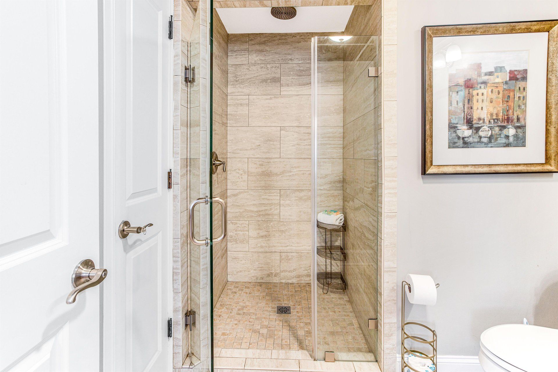 Bathroom with glass shower, beige tile, art print, and white door.
