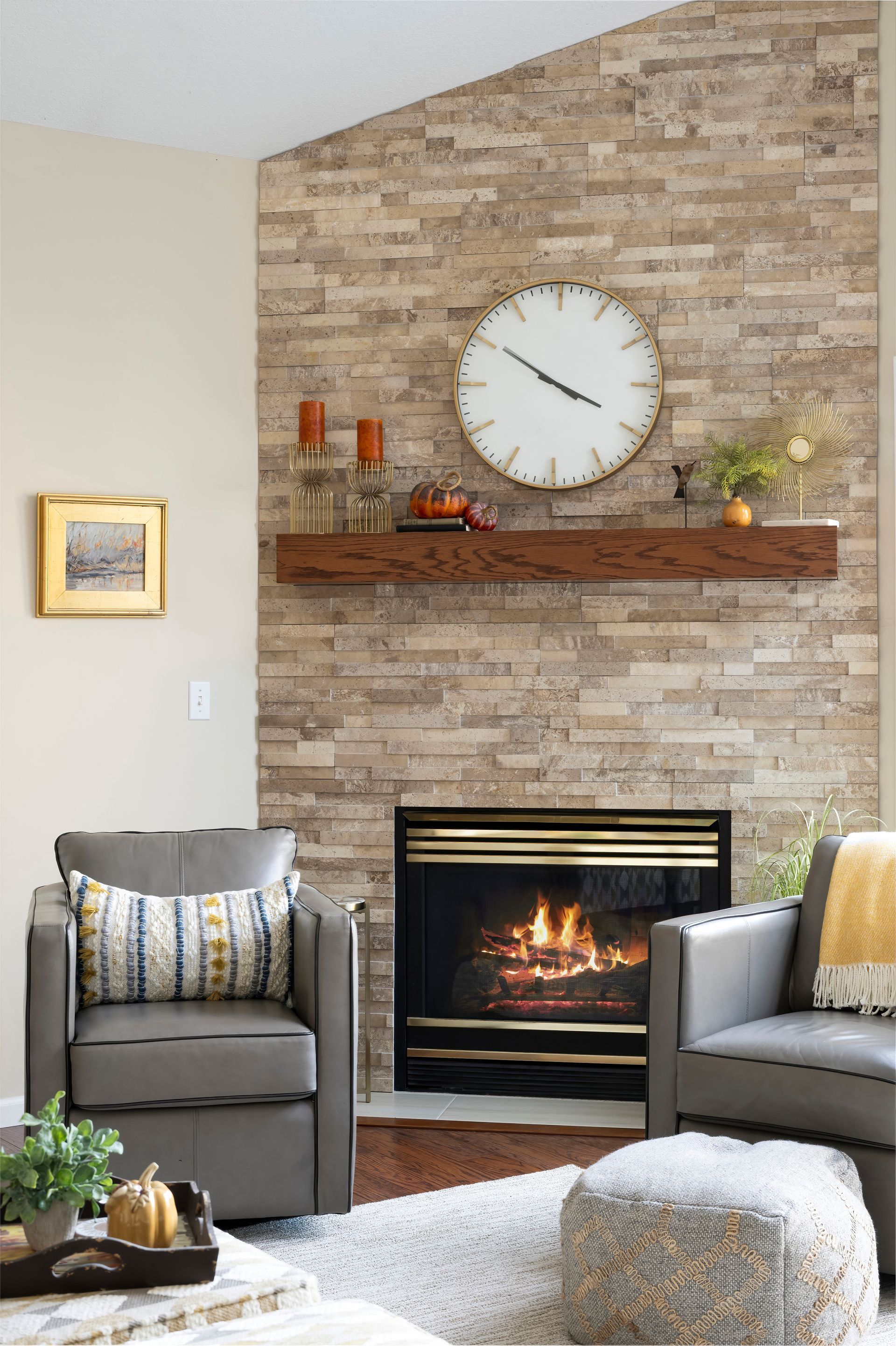 Fireplace with stone surround, wood mantle, clock, candles, and two gray armchairs.