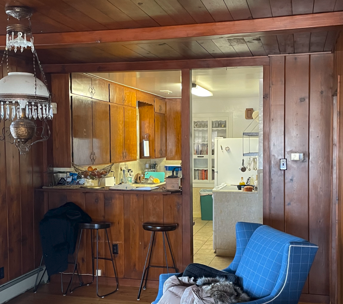 Interior view of a kitchen, seen through an opening in wood-paneled walls. 