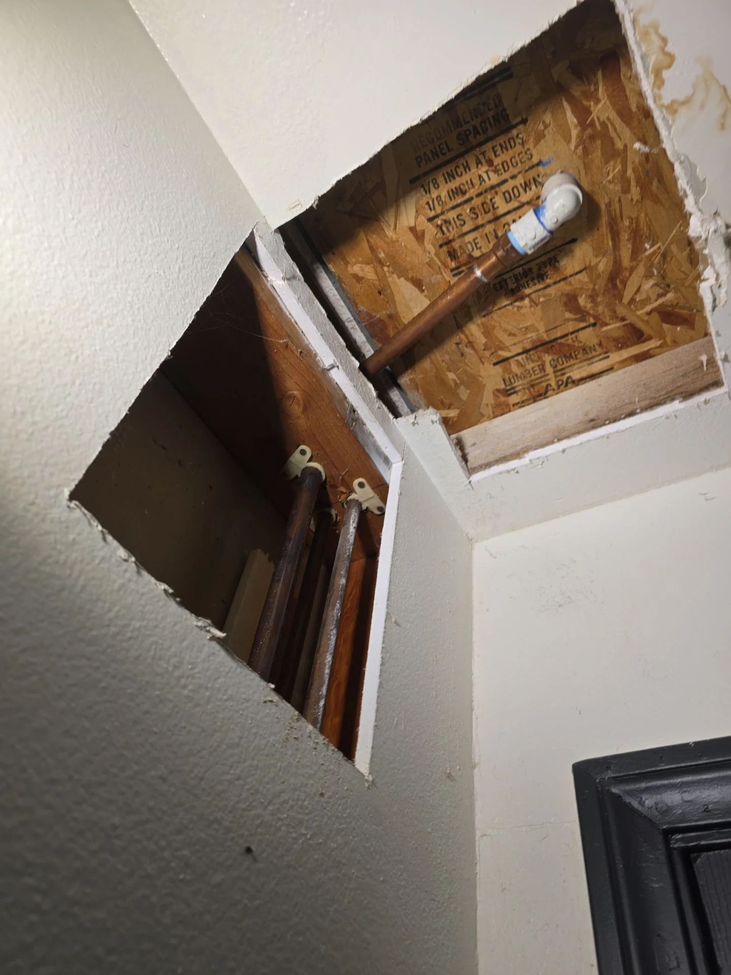 Two rectangular holes cut into a white drywall ceiling and wall, exposing copper plumbing pipes and wooden framing.