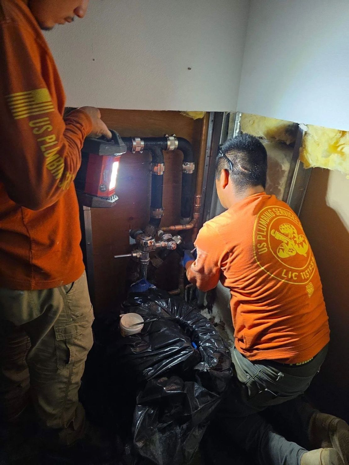 Two people wearing orange work shirts repair pipes in a dimly lit, enclosed crawl space.