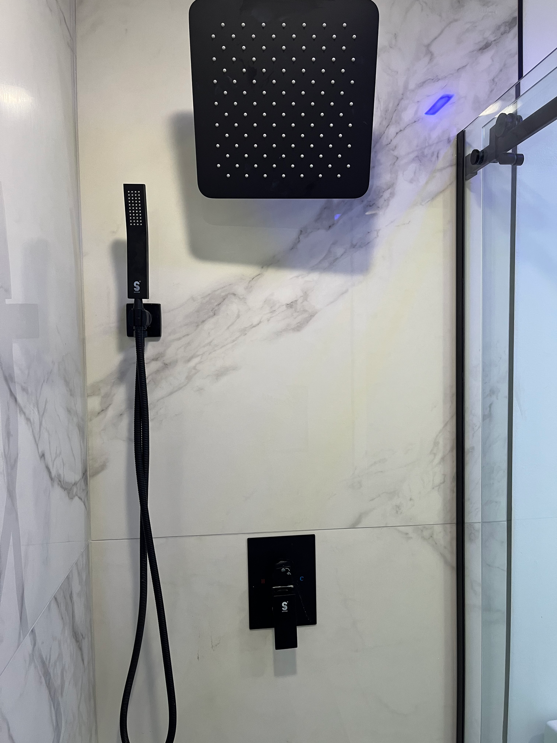 A matte black square rainfall showerhead and a matching handheld shower wand mounted on a white marble-tiled shower wall.