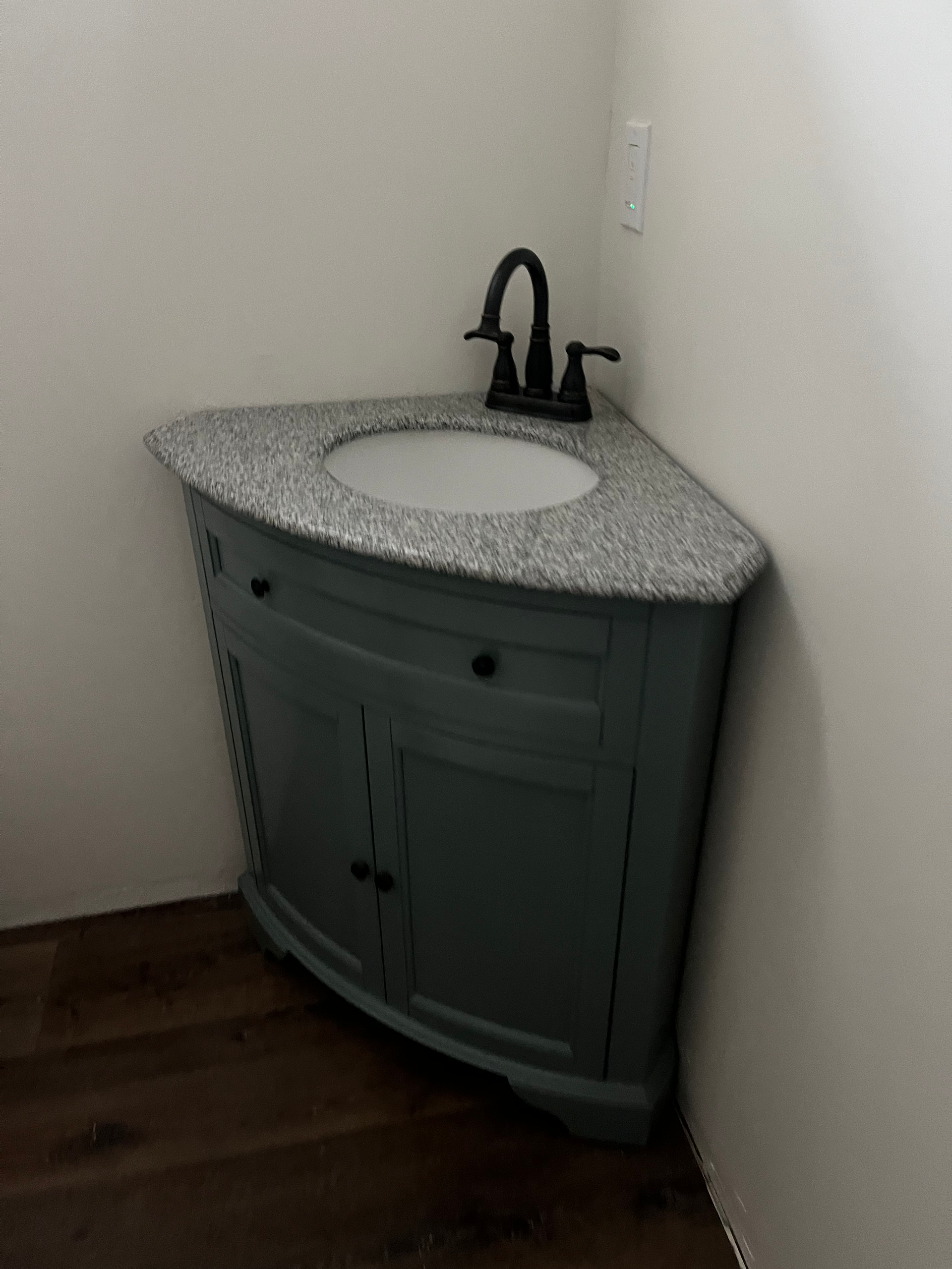 A gray, corner bathroom vanity with a speckled stone countertop and a dark faucet set against white walls.