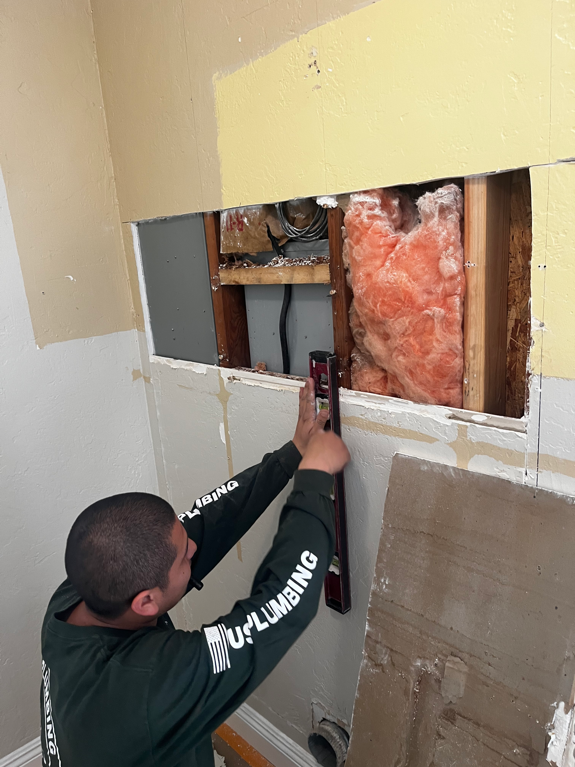 A plumber uses a level to inspect a wall cavity opened during a repair project, revealing wooden framing and insulation.