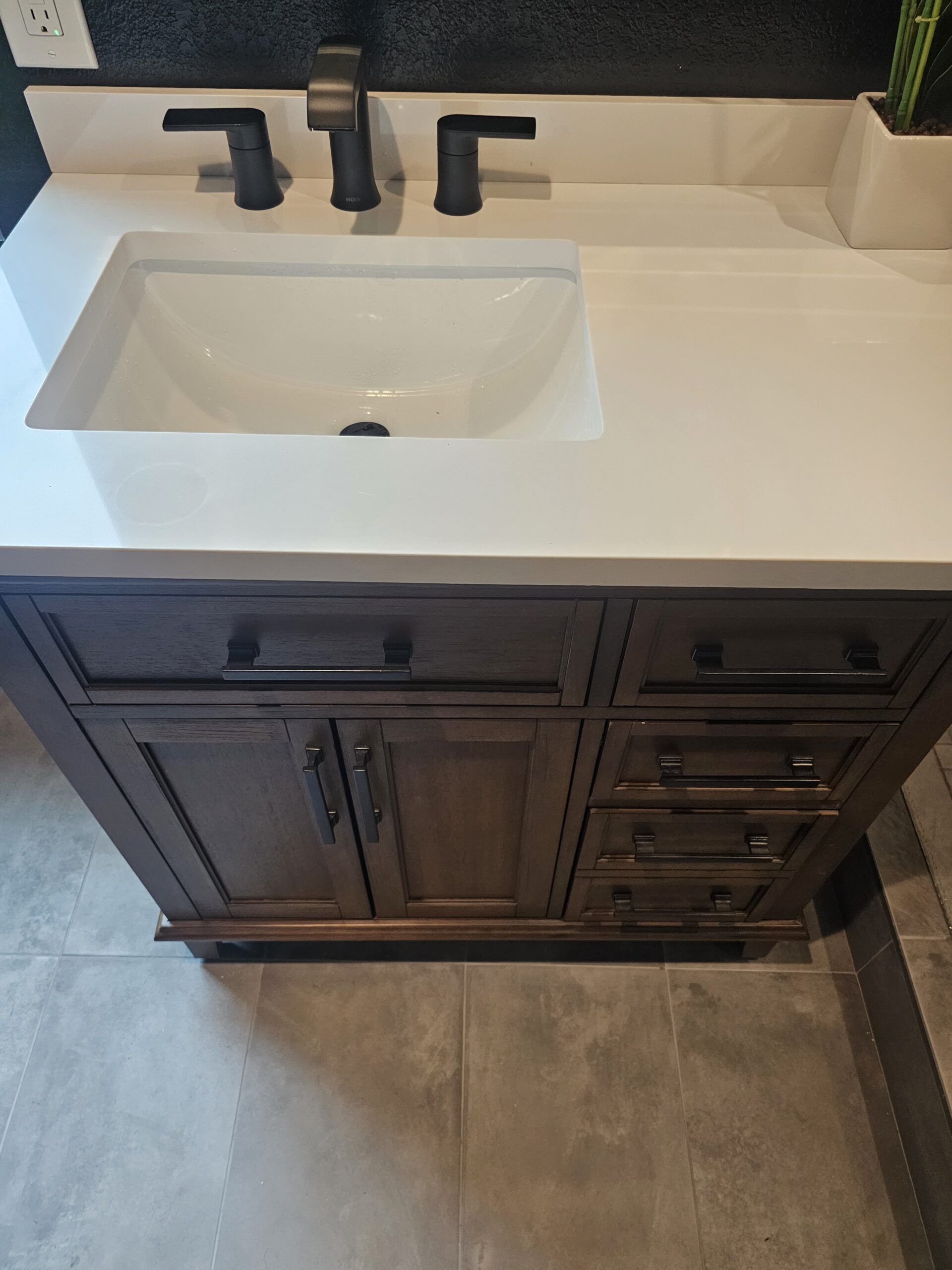 A dark wood bathroom vanity with a white countertop, an undermount sink, and matte black hardware on tile flooring.