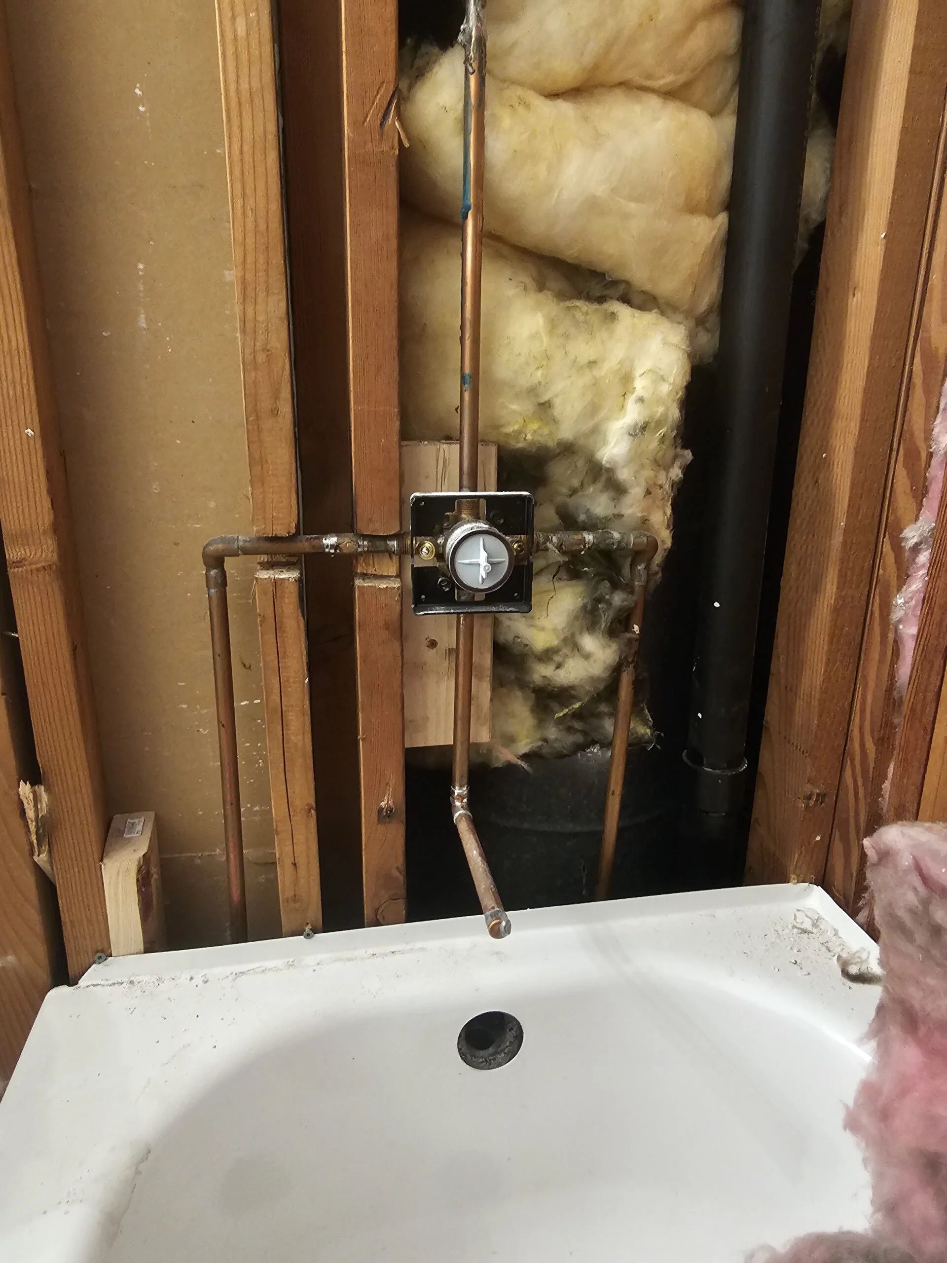 A shower valve and copper pipes are installed within an open wall frame above a bathtub basin.