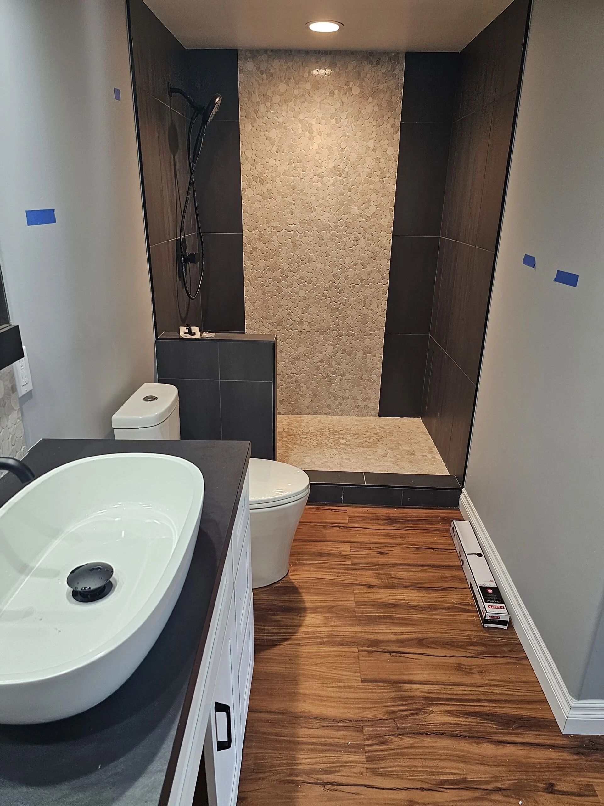 A bathroom renovation showing a walk-in shower with stone tile, dark wall panels, a vanity with a basin, and wood flooring.