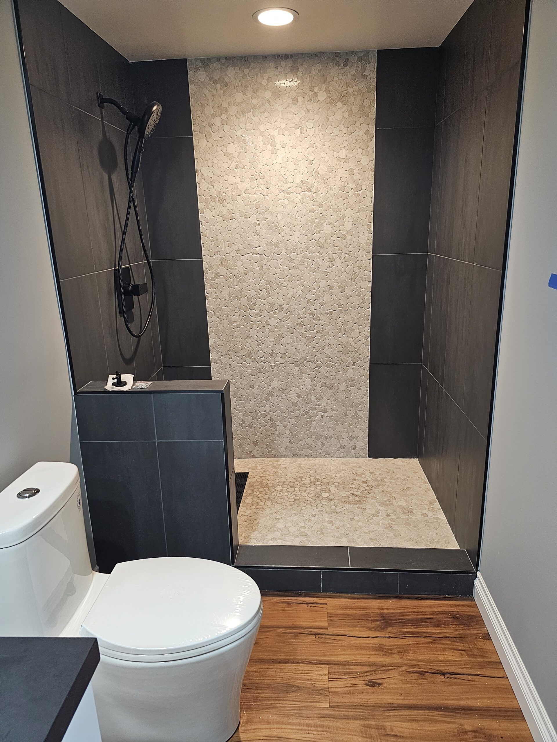 A bathroom featuring a walk-in shower with light stone tile, dark gray wall panels, and a white toilet on wood-look flooring.