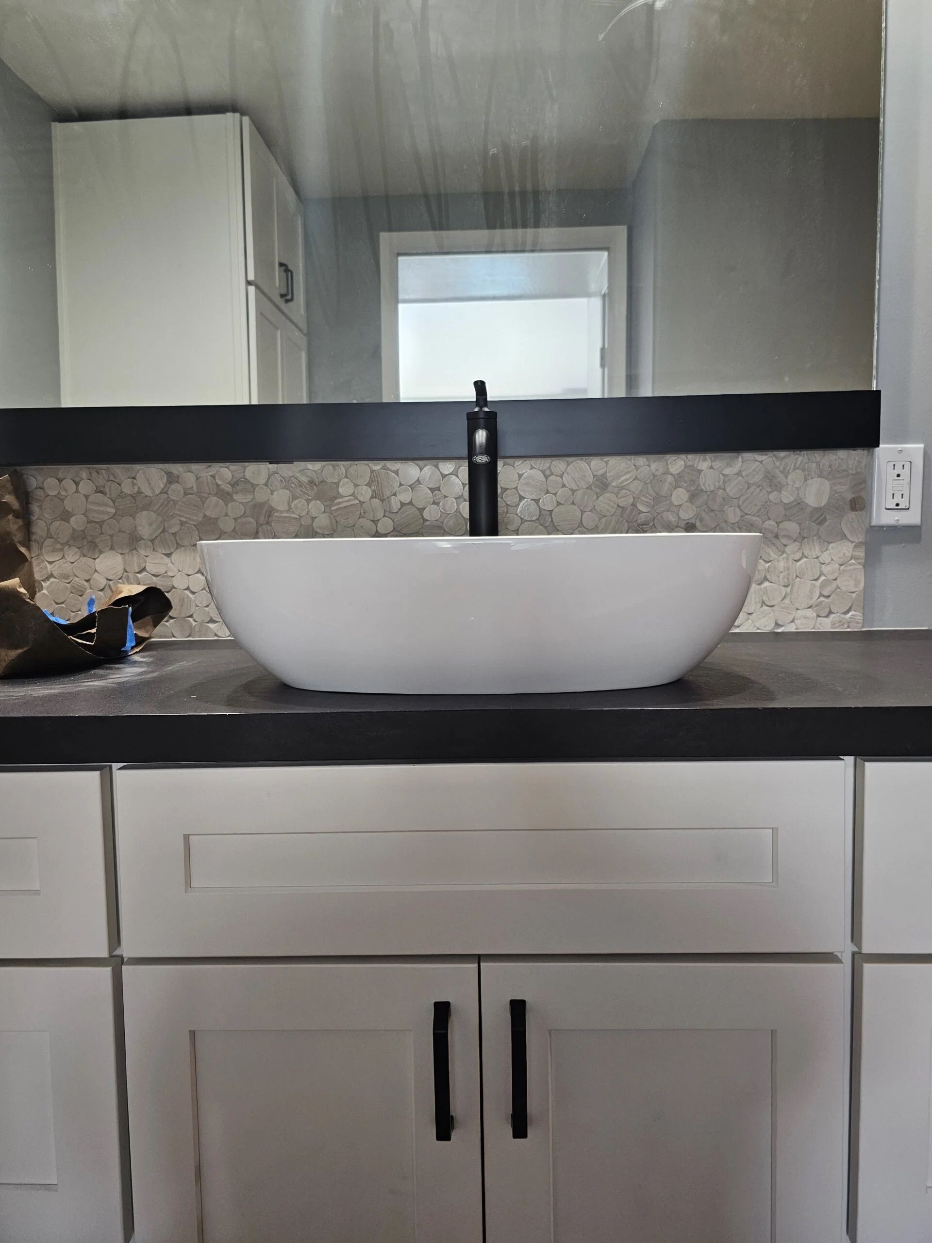 A white vessel sink sits on a dark countertop above white cabinets, centered against a tiled backsplash and mirror.