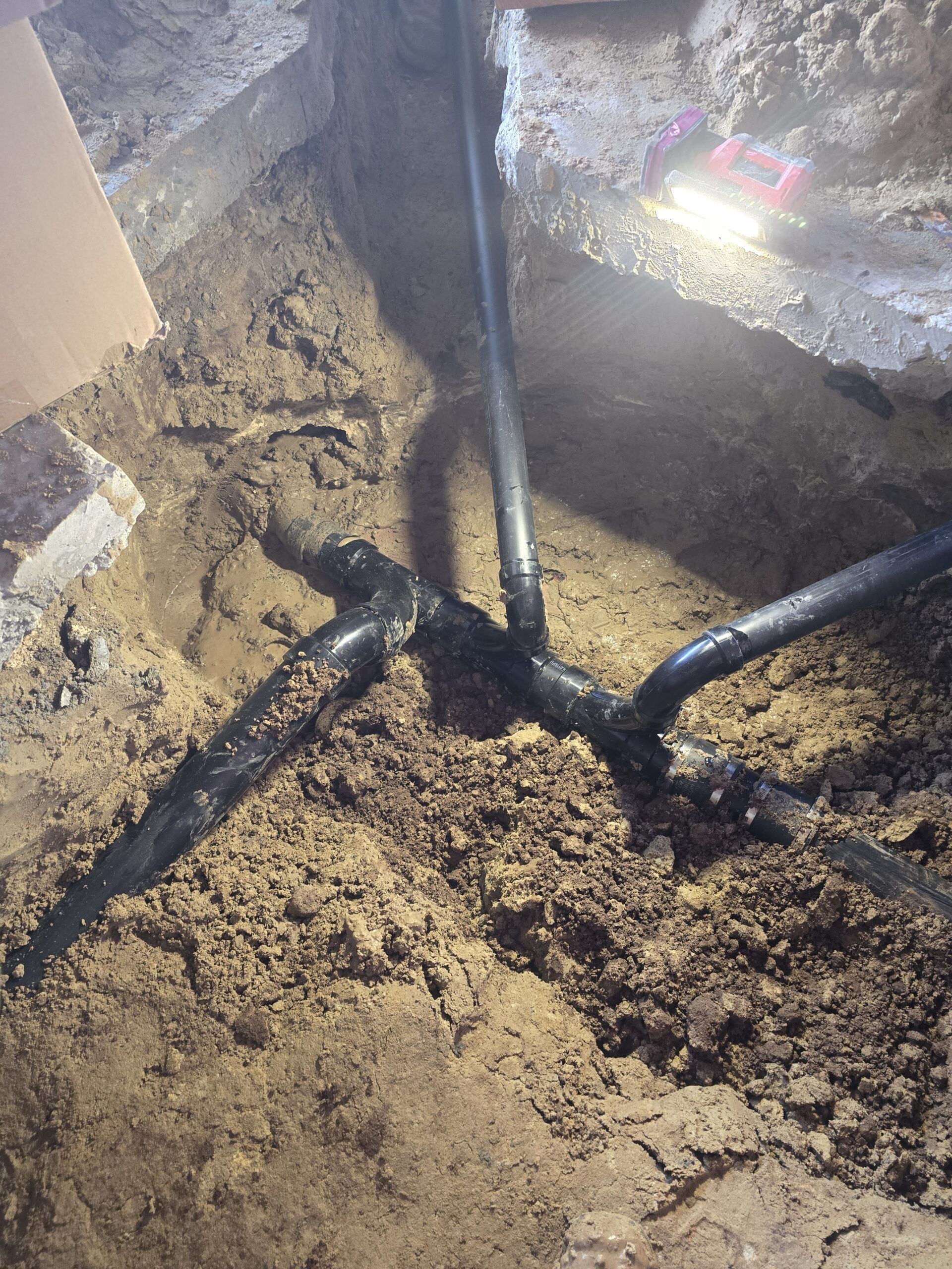 A view into a trench showing black ABS plumbing pipes with a T-junction and elbow connections buried in soil.